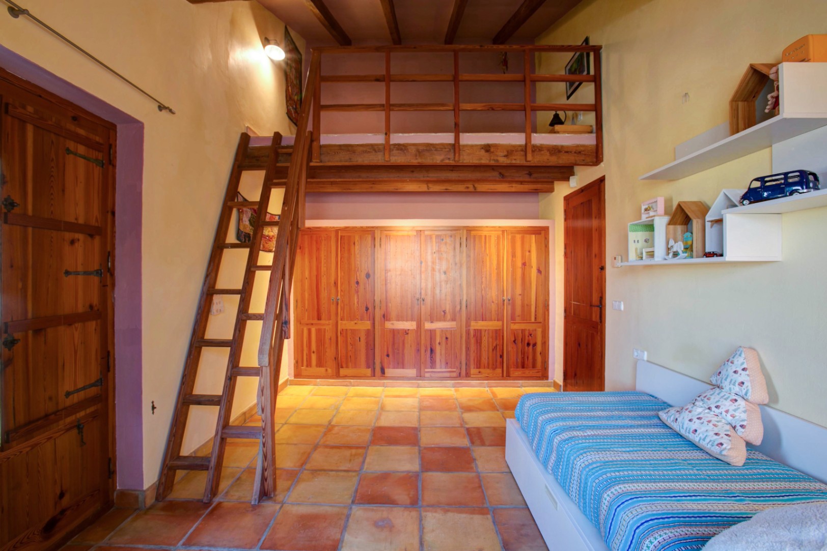 Schlafzimmer mit hölzernem Zwischengeschoss und Leiter, Einbauschränken aus Mobila-Holz und traditionellem Terrakottaboden unter Sichtbalken.