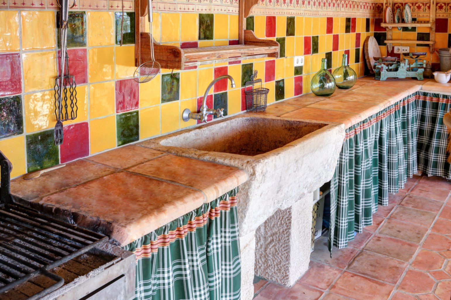 Sommerküche im Freien mit massivem Steinwaschbecken, Arbeitsplatten aus Terrakottafliesen und buntem Fliesenspiegel. Ausgestattet mit Grillbereich und traditionellen Stoffvorhängen.