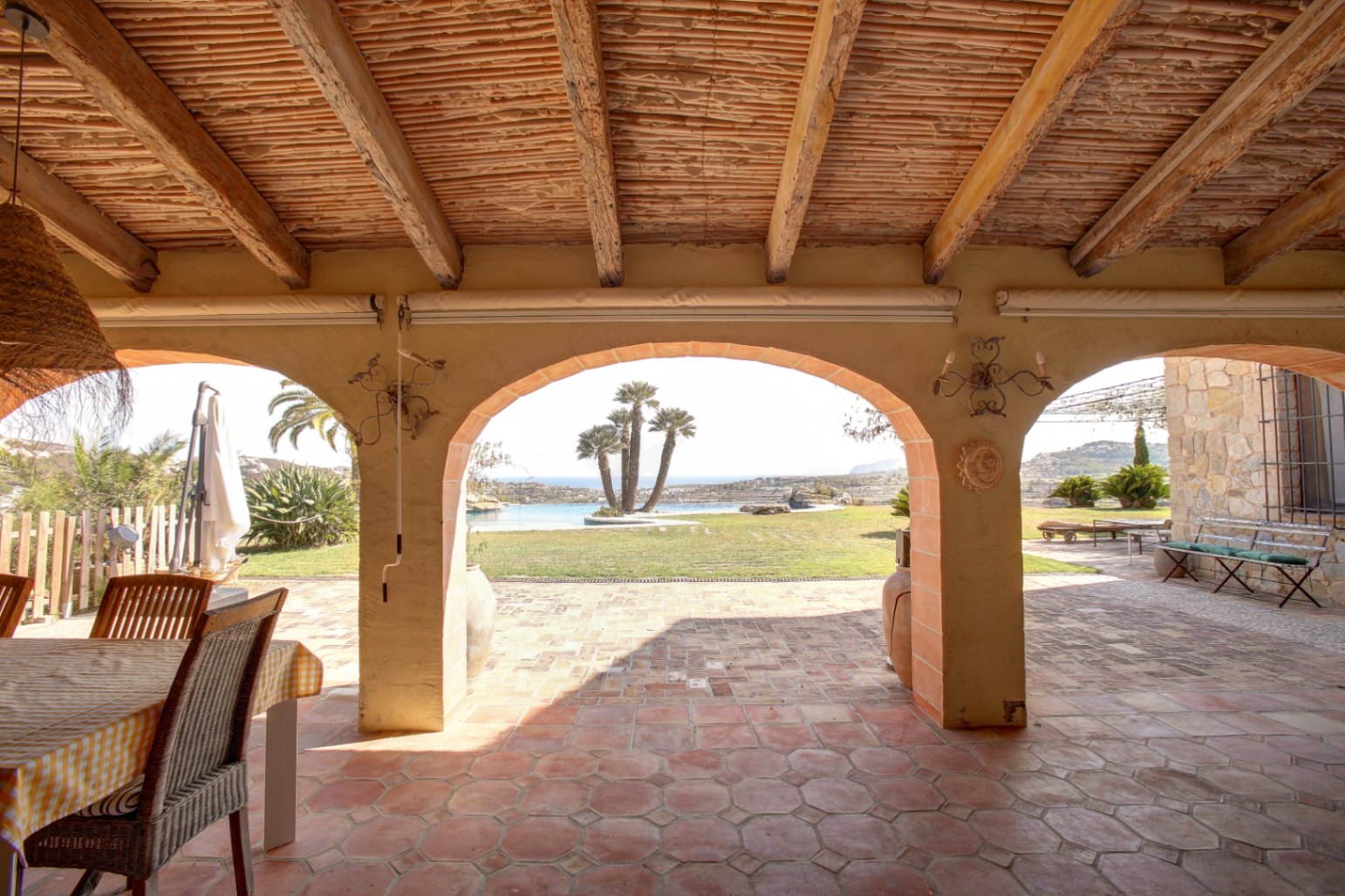 Überdachte Terrasse mit Holzbalken, Schilfrohrdecke und Terrakottaboden. Rundbögen bieten Ausblick auf den Garten, den Pool mit Palmen und das Mittelmeer.