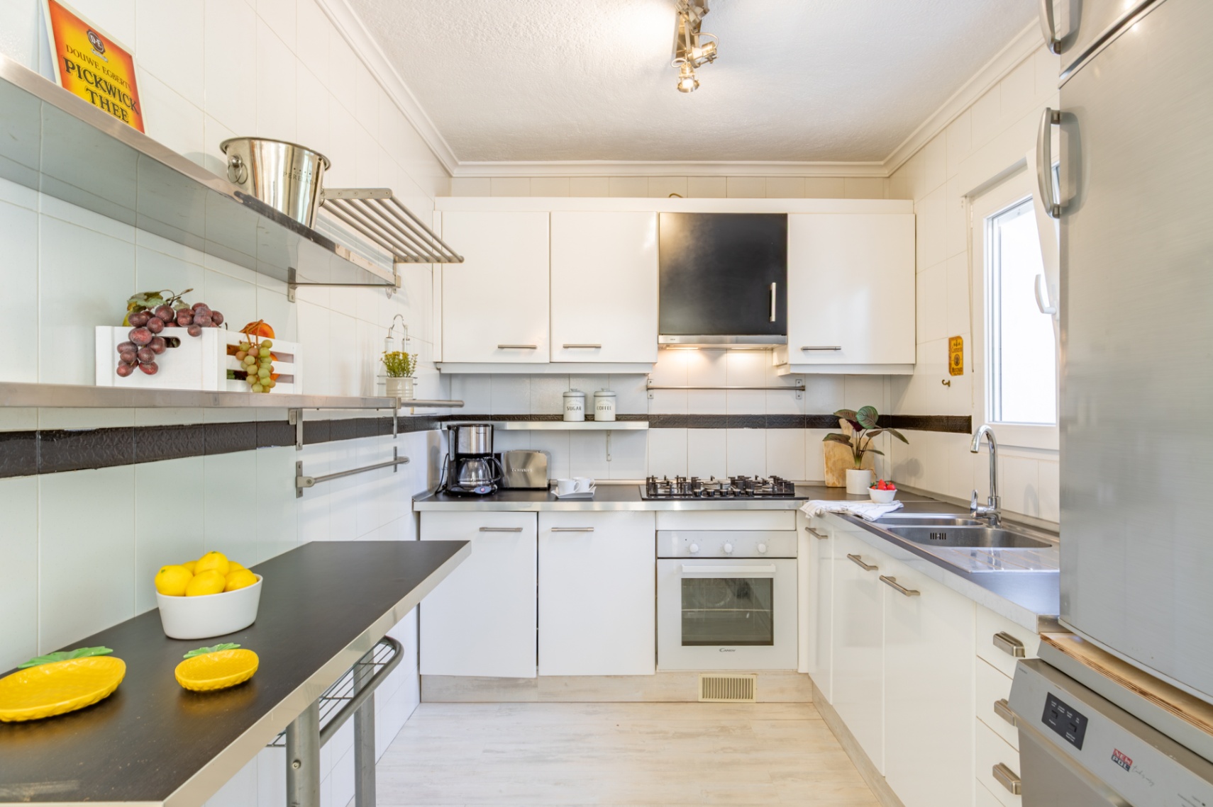 Ausgestattete Küche mit weißen Schränken, dunklen Arbeitsplatten, Edelstahlregalen und Tageslicht. Inklusive Gaskochfeld, Backofen und Doppelspüle aus Edelstahl.