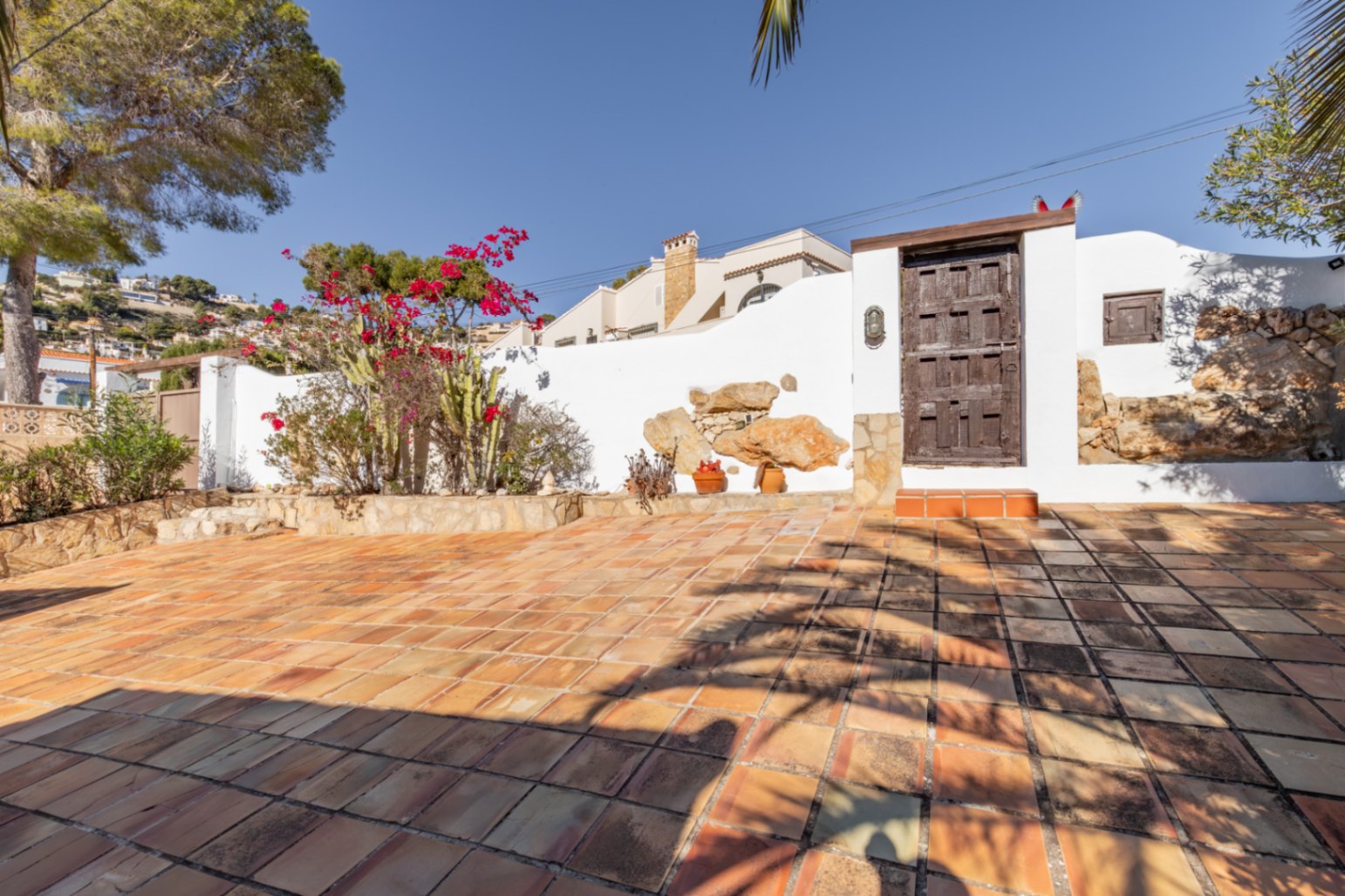 Außen-Terrasse mit Terrakotta-Fliesen, weißen Mauern mit Naturstein-Akzenten und einem rustikalen Holztor, umgeben von mediterranen Bougainvilleen und Kakteen.