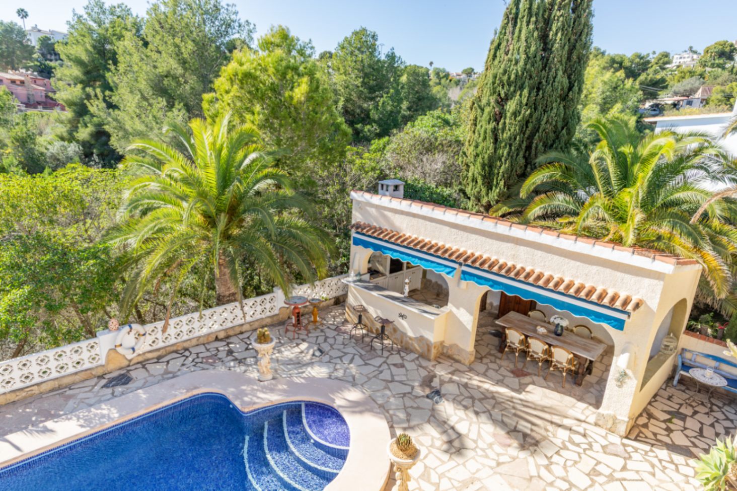 Außenbereich mit nierenförmigem Mosaikpool, Naturstein-Terrasse und überdachter Außenküche, eingebettet in einen Garten mit Palmen und angrenzender Grünzone.