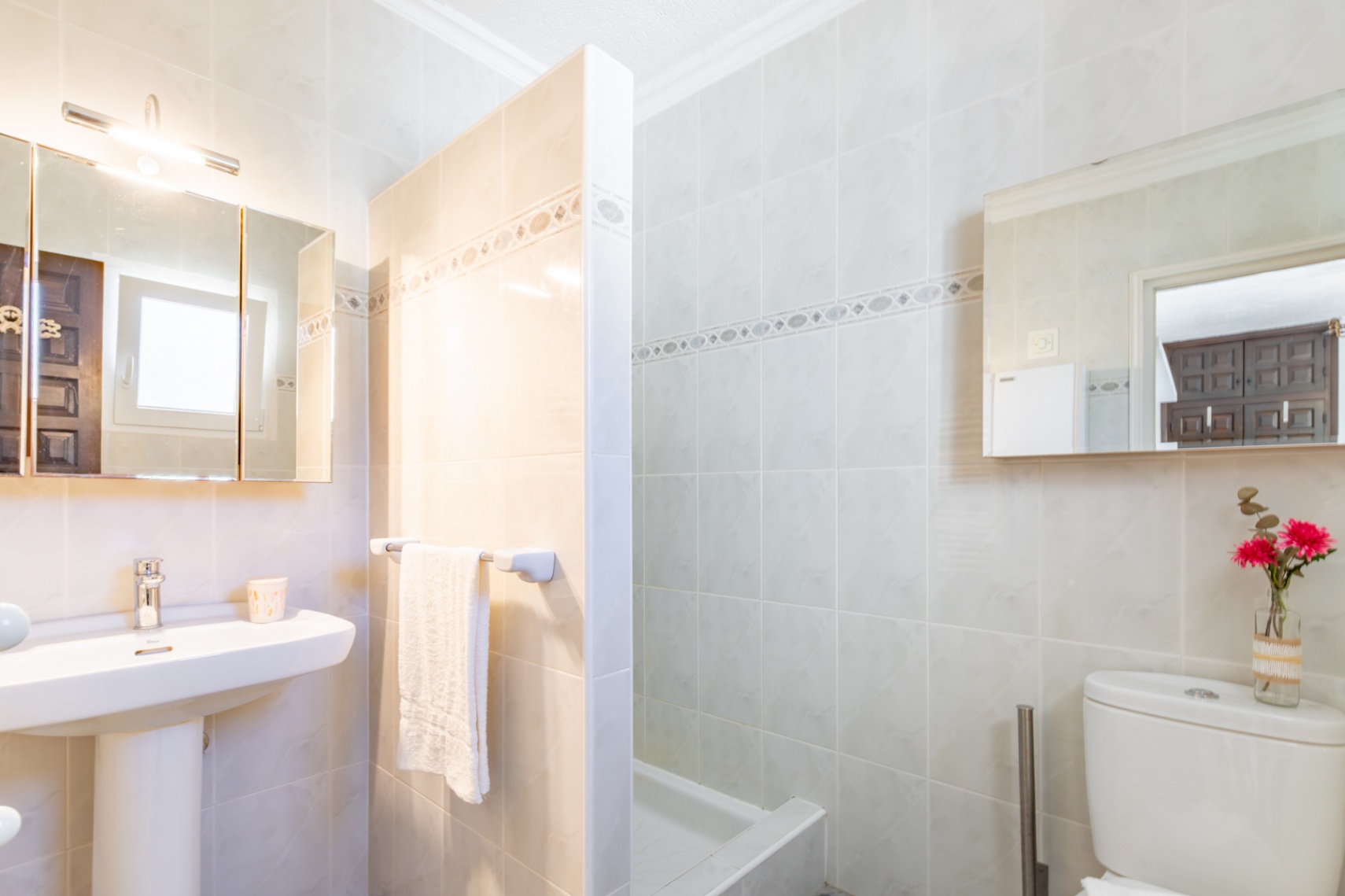 Badezimmer mit cremefarbenen Fliesen, Säulenwaschbecken, Spiegelschrank mit Beleuchtung und einer durch eine Trennwand abgeteilten Dusche.