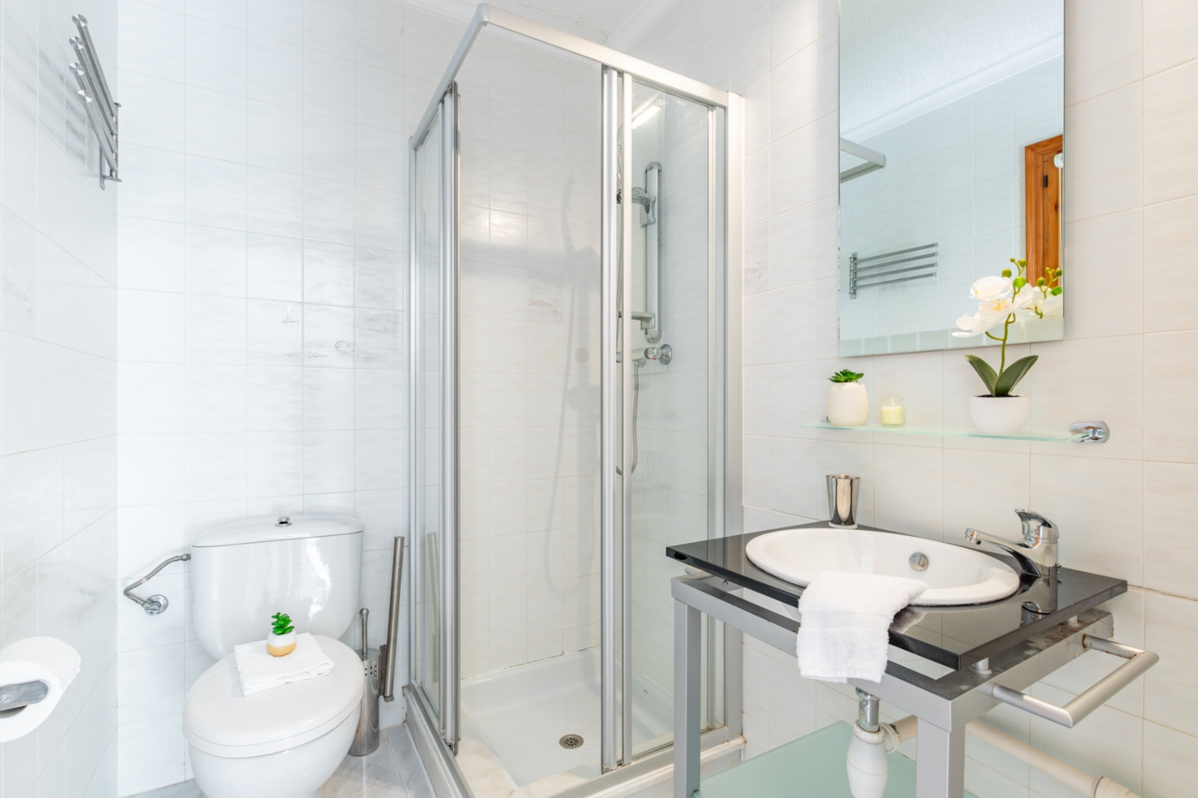 Badezimmer mit weiß gefliesten Wänden, einer gläsernen Eckduschkabine und einem Waschtisch mit dunkler Steinplatte und integriertem Becken.