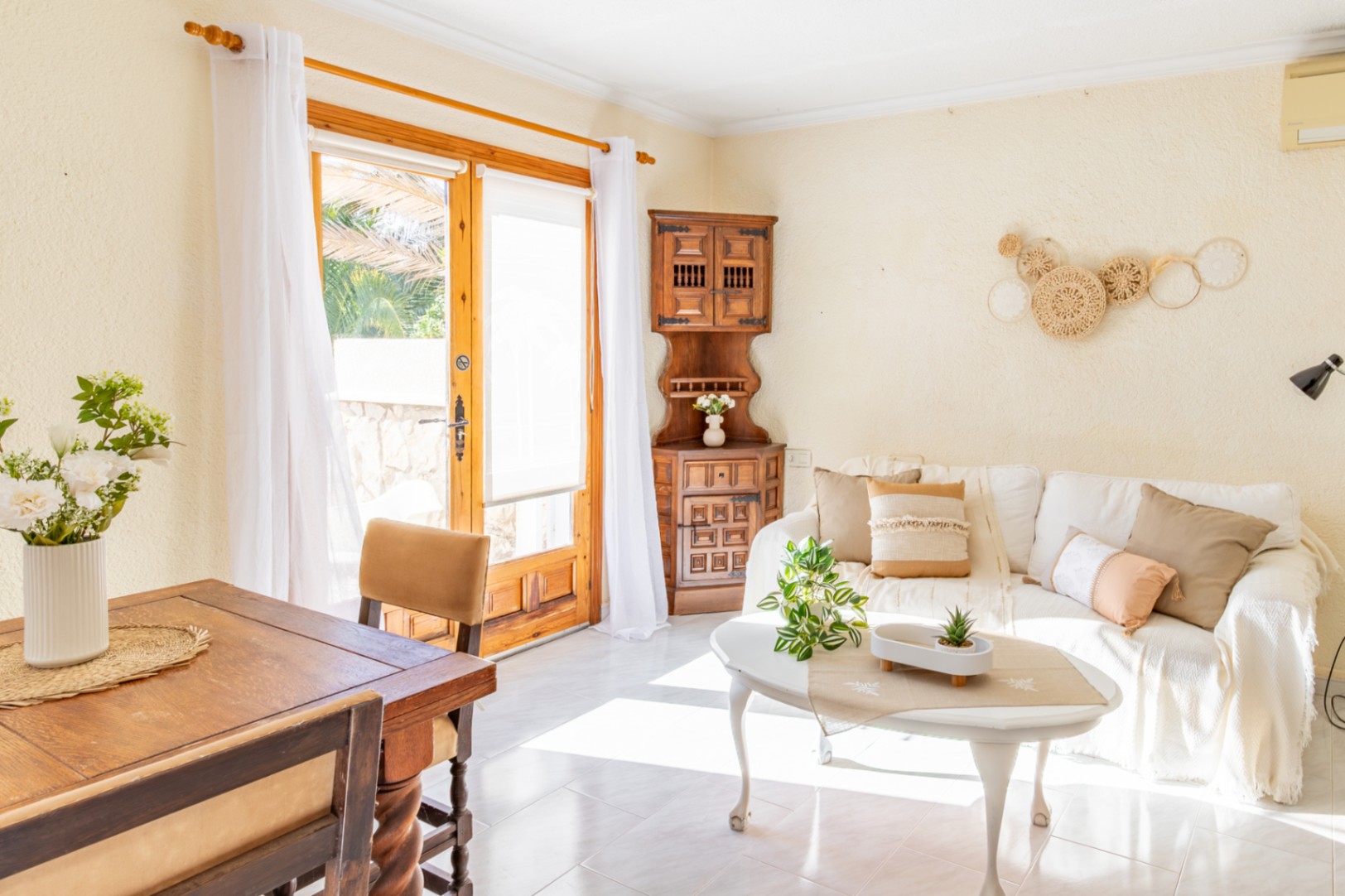 Heller Wohn- und Essbereich mit rustikalen Holzmöbeln, weißem Sofa und direktem Zugang zum Außenbereich durch eine große Glastür mit Naturholzrahmen.