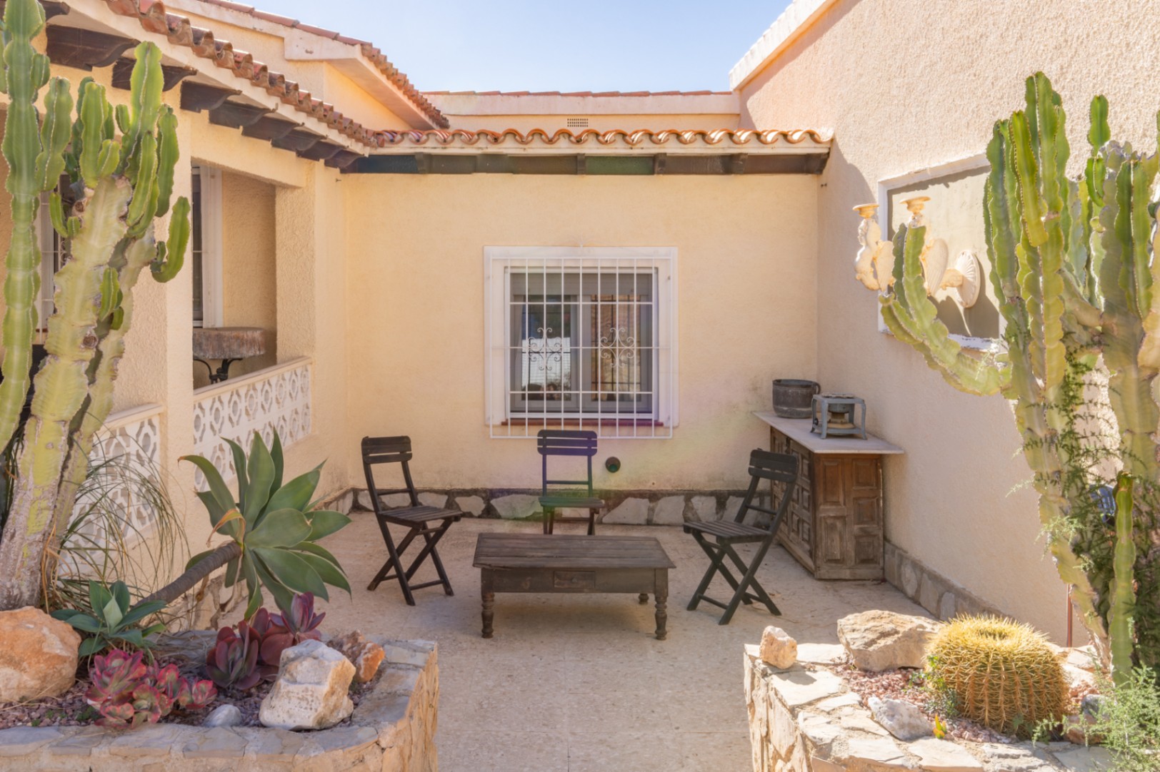 Mediterraner Außenhof mit Steinboden, rustikalen Holzmöbeln und gemauerten Pflanzgefäßen mit Kakteen und Sukkulenten unter der Sonne der Costa Blanca.