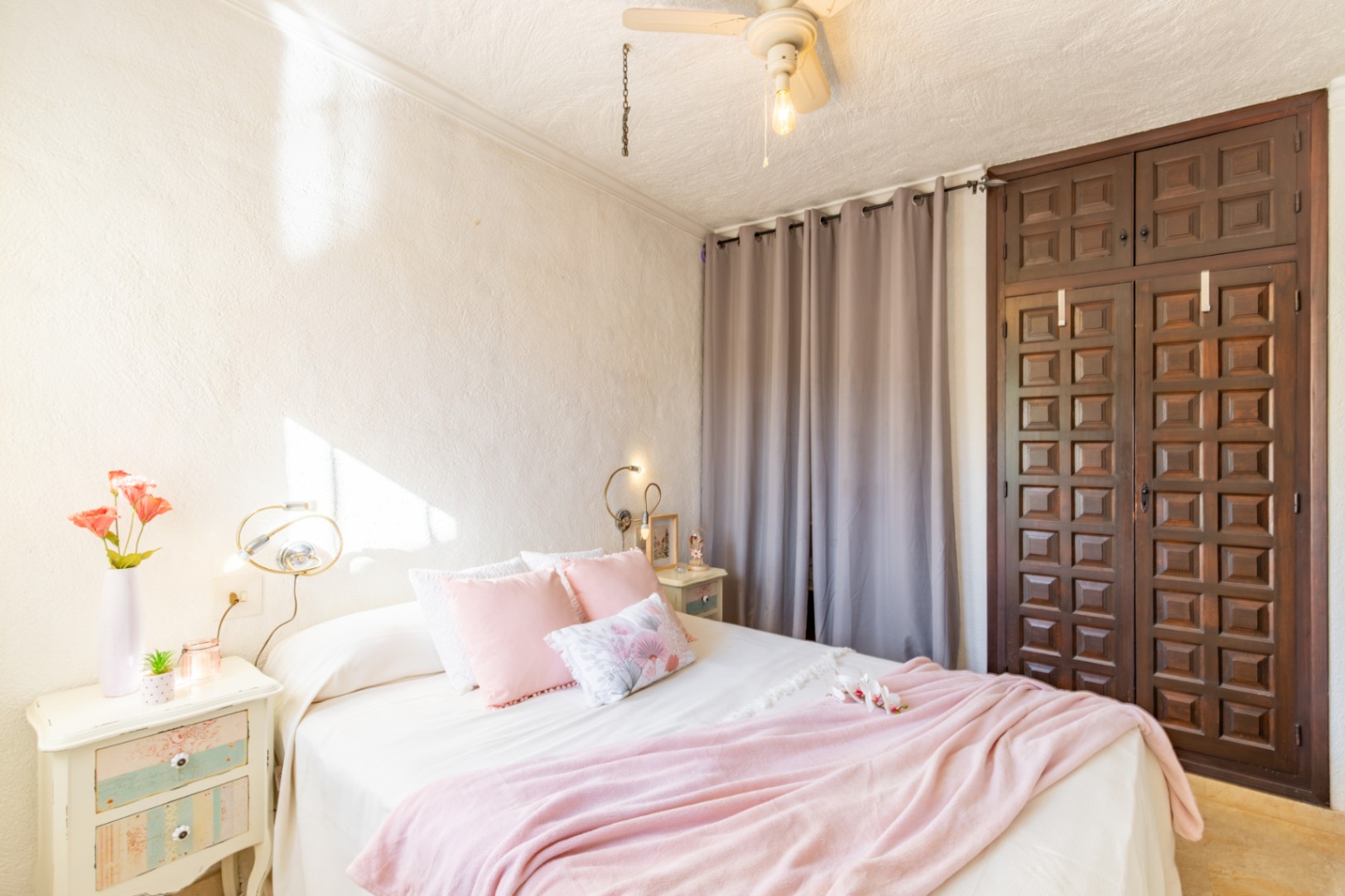 Schlafzimmer mit dunklem Einbauschrank aus Holz, strukturierten weißen Wänden und Deckenventilator. Der Raum wird durch seitliches Tageslicht hell beleuchtet.