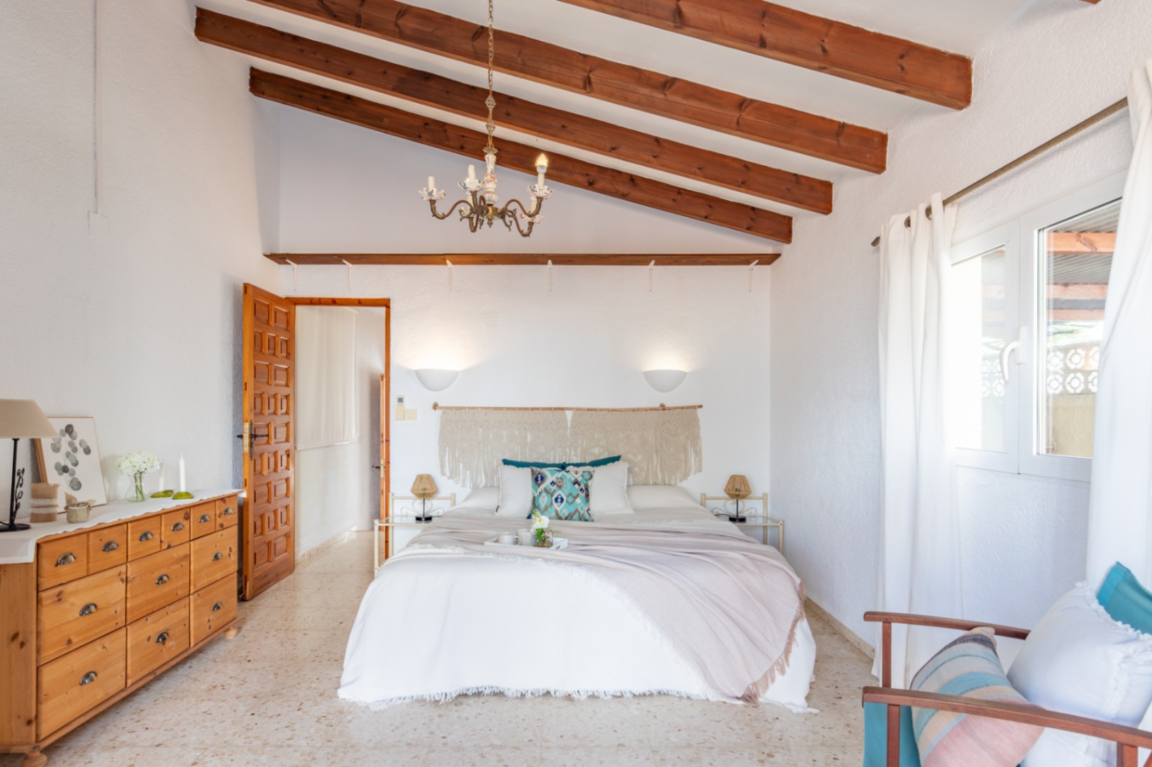 Schlafzimmer mit hohen Decken und sichtbaren Holzbalken, Terrazzoboden und mediterraner Dekoration mit Makramee-Kopfteil und Naturholzmöbeln.