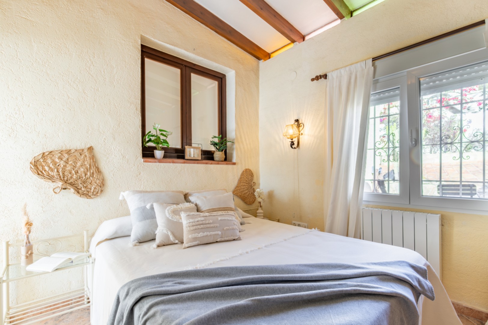 Schlafzimmer mit sichtbaren Holzbalken an der Decke, rustikal strukturierten Wänden und zwei Fenstern. Die Einrichtung besticht durch mediterrane Details.