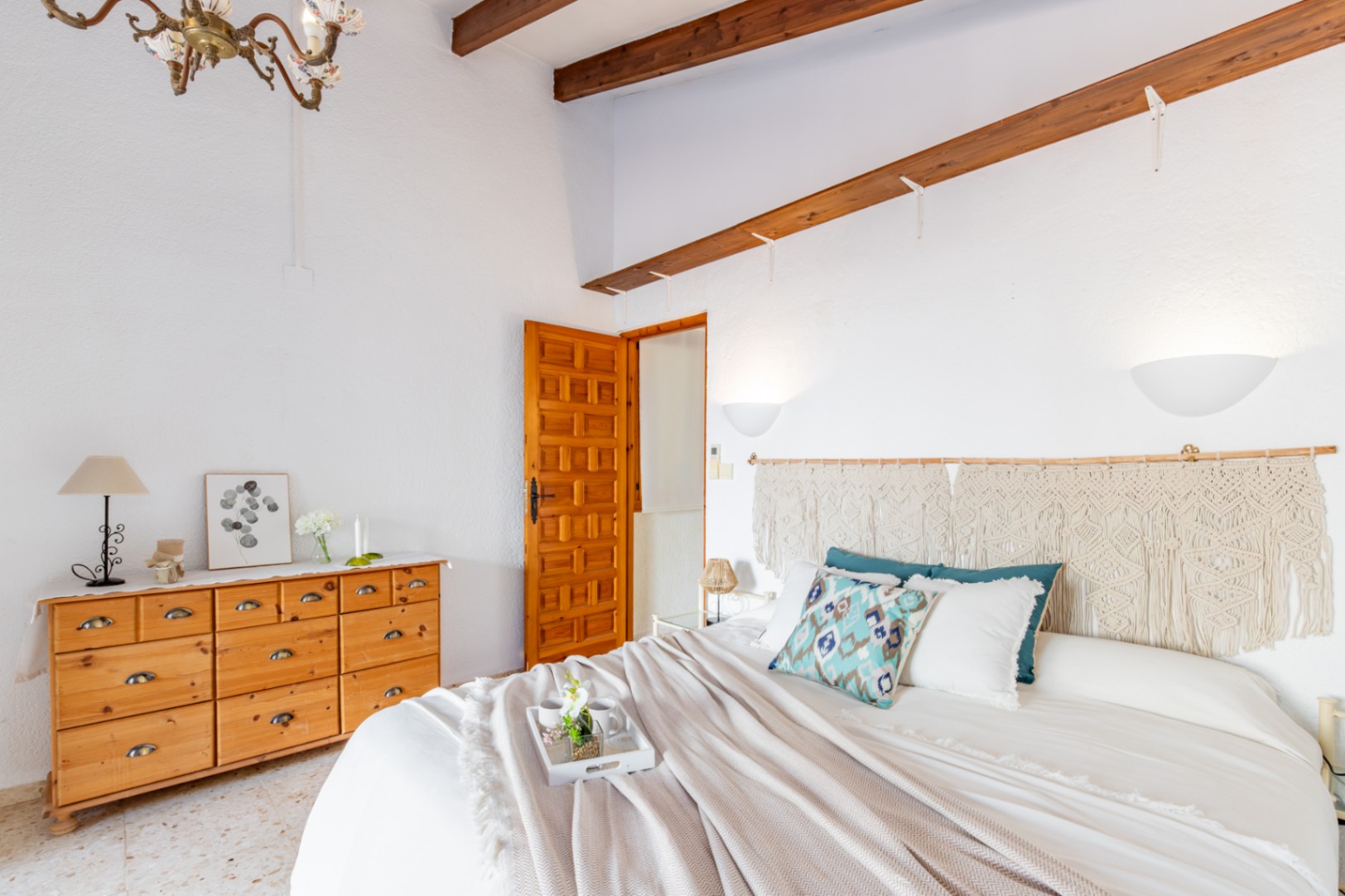 Schlafzimmer mit sichtbaren Holzbalken, einer Kiefernkommode und einem Makramee-Kopfteil. Ausgestattet mit originalem Terrazzoboden und einer traditionell geschnitzten Holztür.