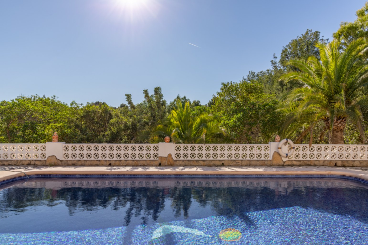 Südlich ausgerichteter Pool mit blauen Mosaikfliesen, begrenzt durch eine weiße Ziermauer und umgeben von üppiger mediterraner Vegetation.