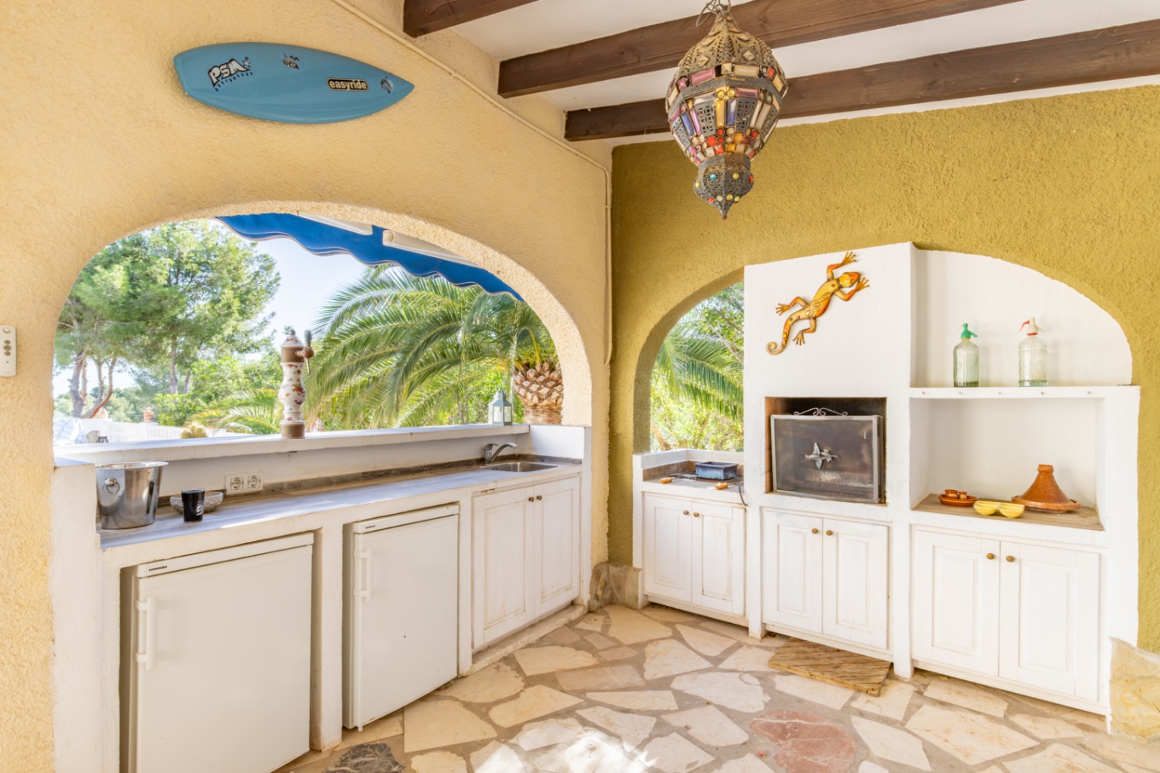 Überdachte Sommerküche mit Holzbalkendecke, gemauertem Grill, mehreren Kühlschränken und Spüle. Der Bereich bietet Natursteinboden und Blick auf die mediterrane Vegetation.