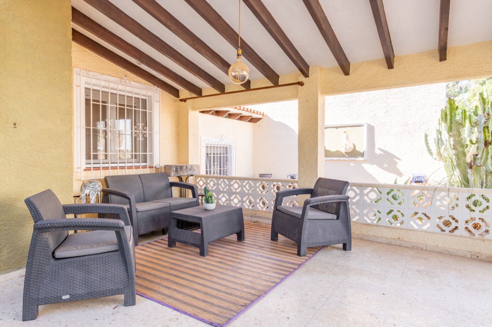 Überdachte Terrasse mit sichtbaren Holzbalken, grauen Gartenmöbeln in Rattan-Optik und einer dekorativen, geometrischen Balustrade.