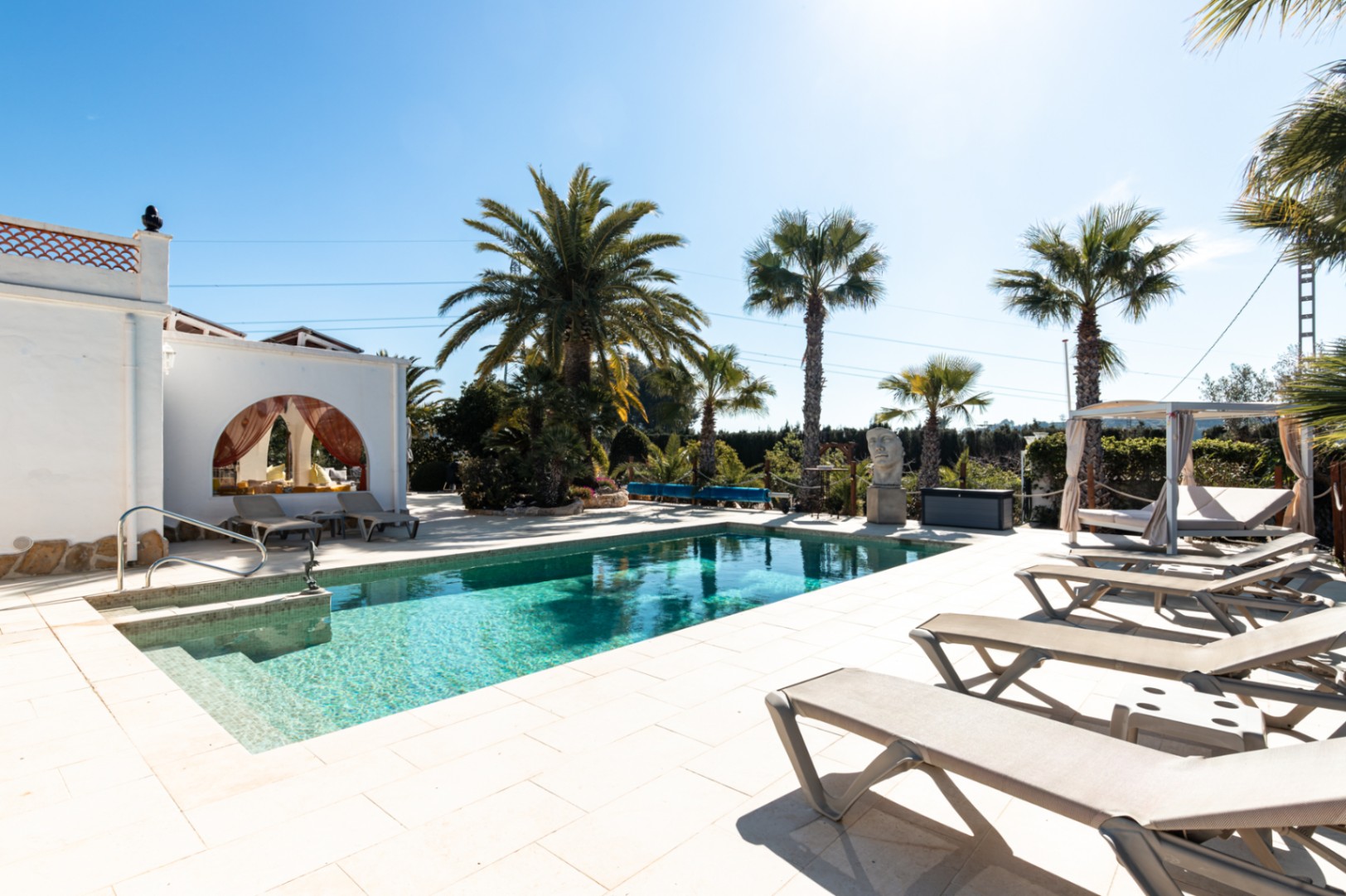 9,5 x 5 m großer Pool mit heller Steinterrasse, Sonnenliegen, balinesischem Bett und mediterranem Garten mit Palmen.