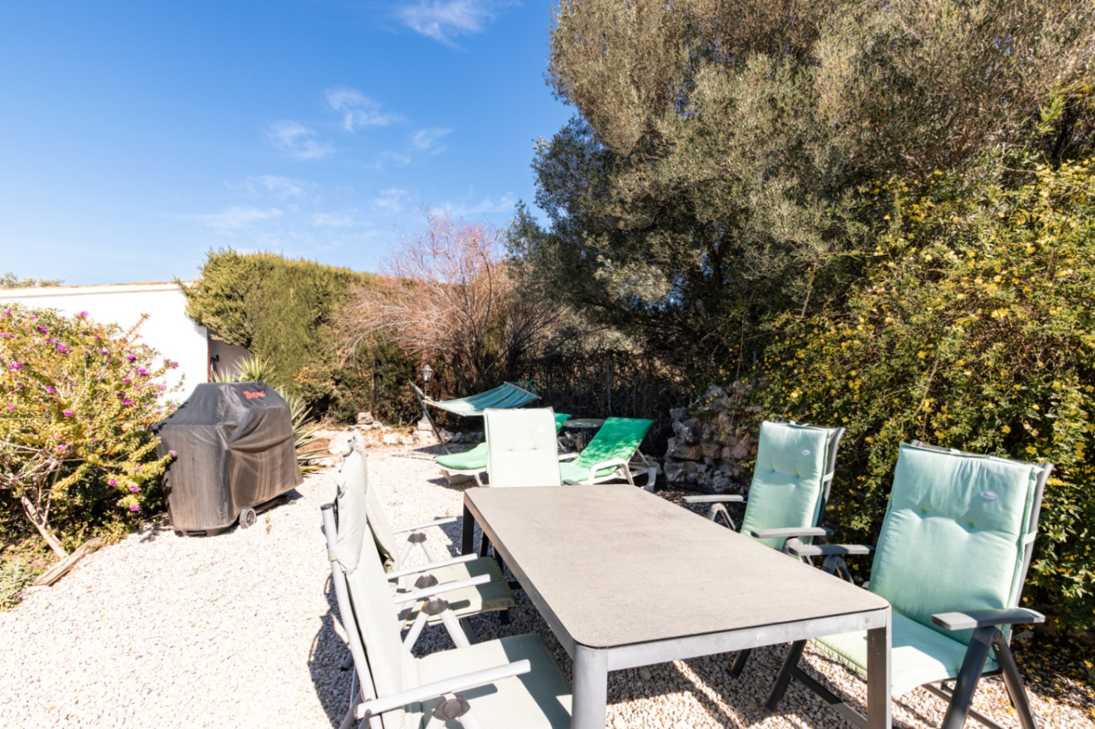 Außen-Essbereich auf Kies mit rechteckigem Tisch, Polsterstühlen, einem abgedeckten Grill und einer Hängematte inmitten mediterraner Bepflanzung.