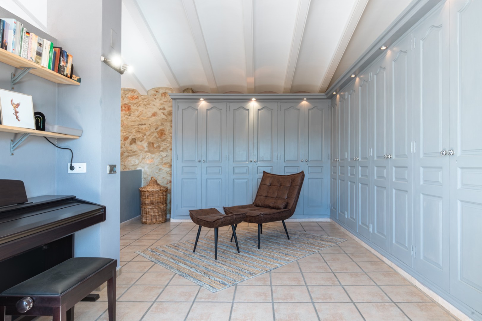 Durchgangszimmer mit großzügigen Einbauschränken in Pastellblau, Natursteinwand, weißen Deckenbalken und einem Entspannungsbereich mit Ledersessel und Klavier.
