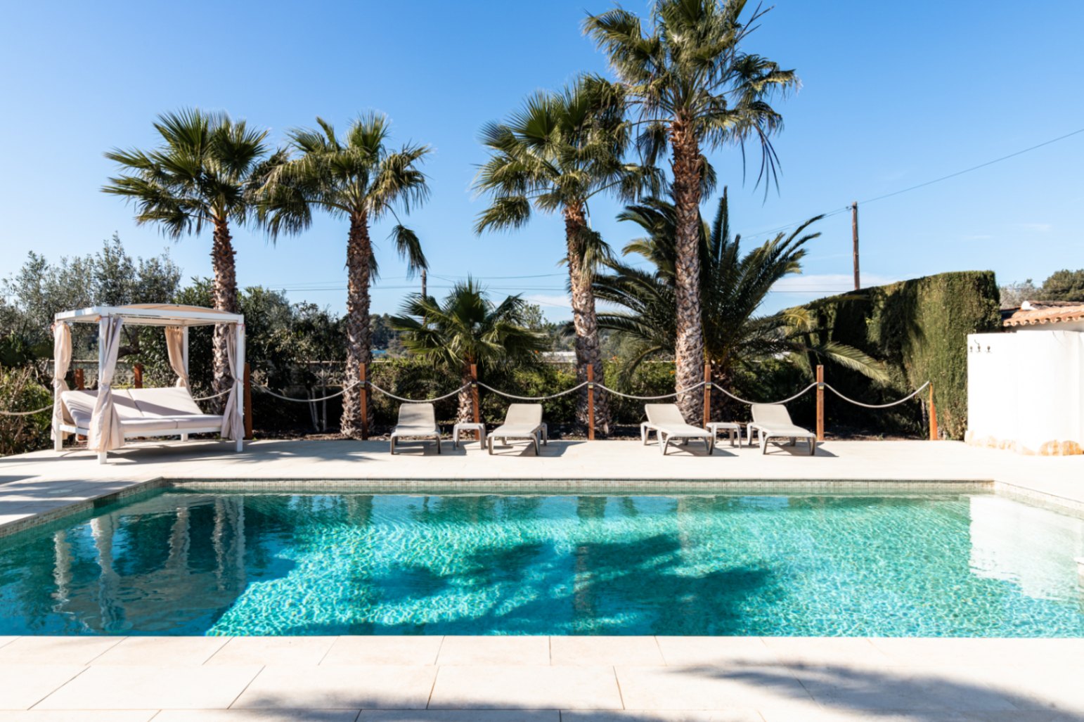 Ein 9,5 x 5 m großer Pool mit gepflasterter Sonnenterrasse, Sonnenliegen, einem Daybed mit Vorhängen und hohen Palmen im Hintergrund.