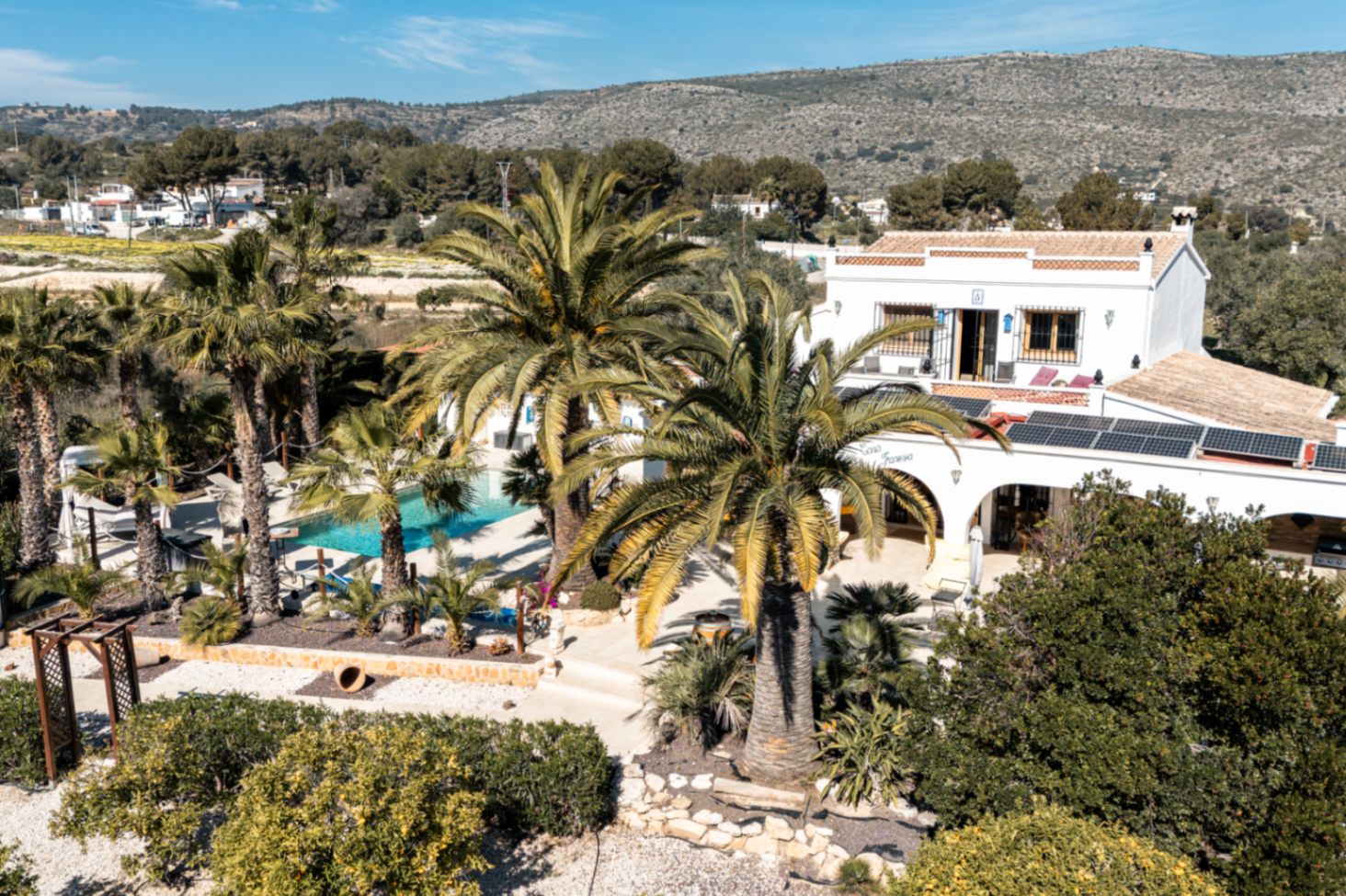 Erhöhte Ansicht der Finca mit Fokus auf die Solaranlage, den von Palmen gesäumten Pool und die traditionelle weiße Architektur vor bergiger Kulisse.