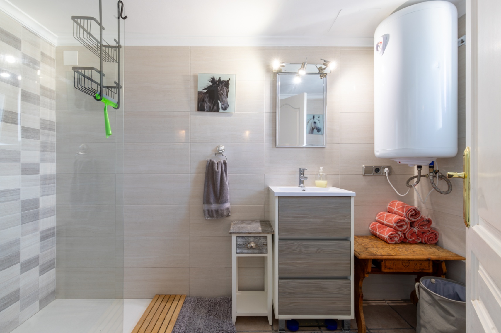 Funktionales Badezimmer mit begehbarer Dusche, Glastrennwand und strukturierten Fliesen. Es bietet einen grauen Waschtisch, Elektro-Warmwasserspeicher und Holzmöbel.