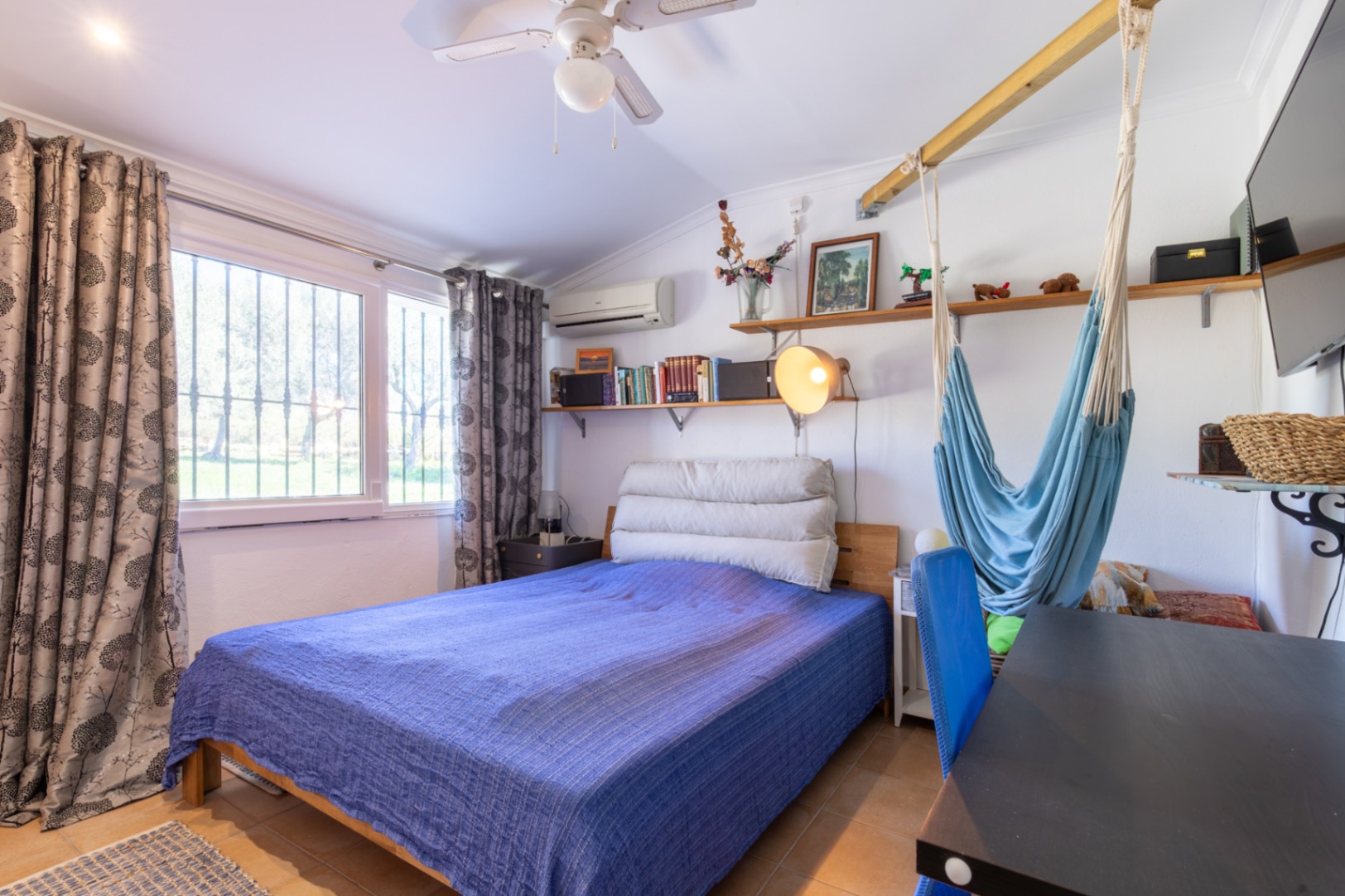 Funktionales Schlafzimmer mit Doppelbett, Schreibtisch, Holzregalen und Hängematte. Mit Klimaanlage, Deckenventilator und viel natürlichem Licht durch das große Fenster.