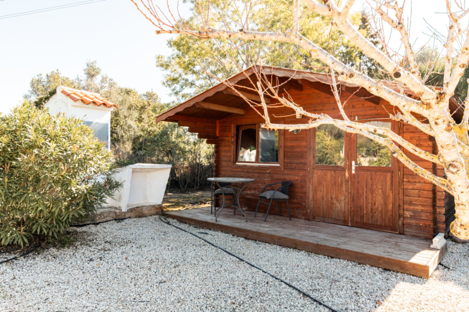 Gartenhaus aus Holz mit kleiner Veranda, Glastüren und Sitzbereich auf einem Kiesgrundstück, umgeben von natürlicher Vegetation.