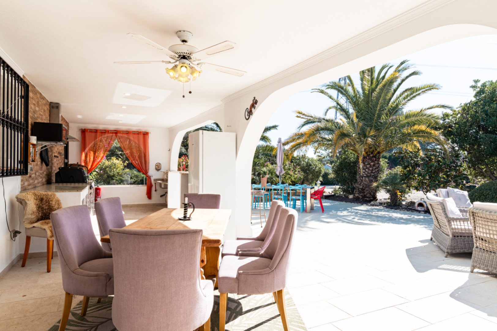 Großzügige überdachte Naya mit Essbereich, Deckenventilator und Außenküche. Die Terrasse bietet direkten Zugang zum sonnigen Garten mit Palmen und Lounge-Möbeln.