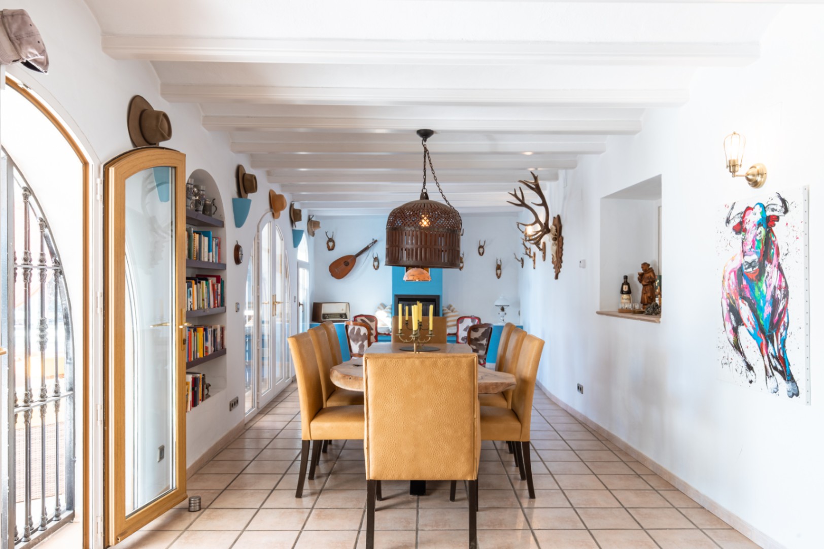 Großzügiges Esszimmer mit weißen Sichtbalken, Massivholztisch und senfgelben Polsterstühlen. Der Raum bietet Rundbogentüren, Einbauregale und eine rustikale, eklektische Dekoration.