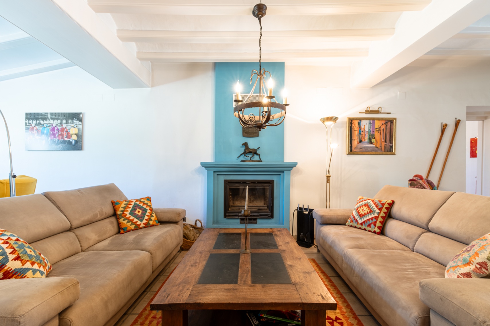 Heller Wohnbereich mit weißer Balkendecke, einem markanten blauen Kamin und gemütlichen Sofas um einen massiven Couchtisch aus Holz und Schiefer.