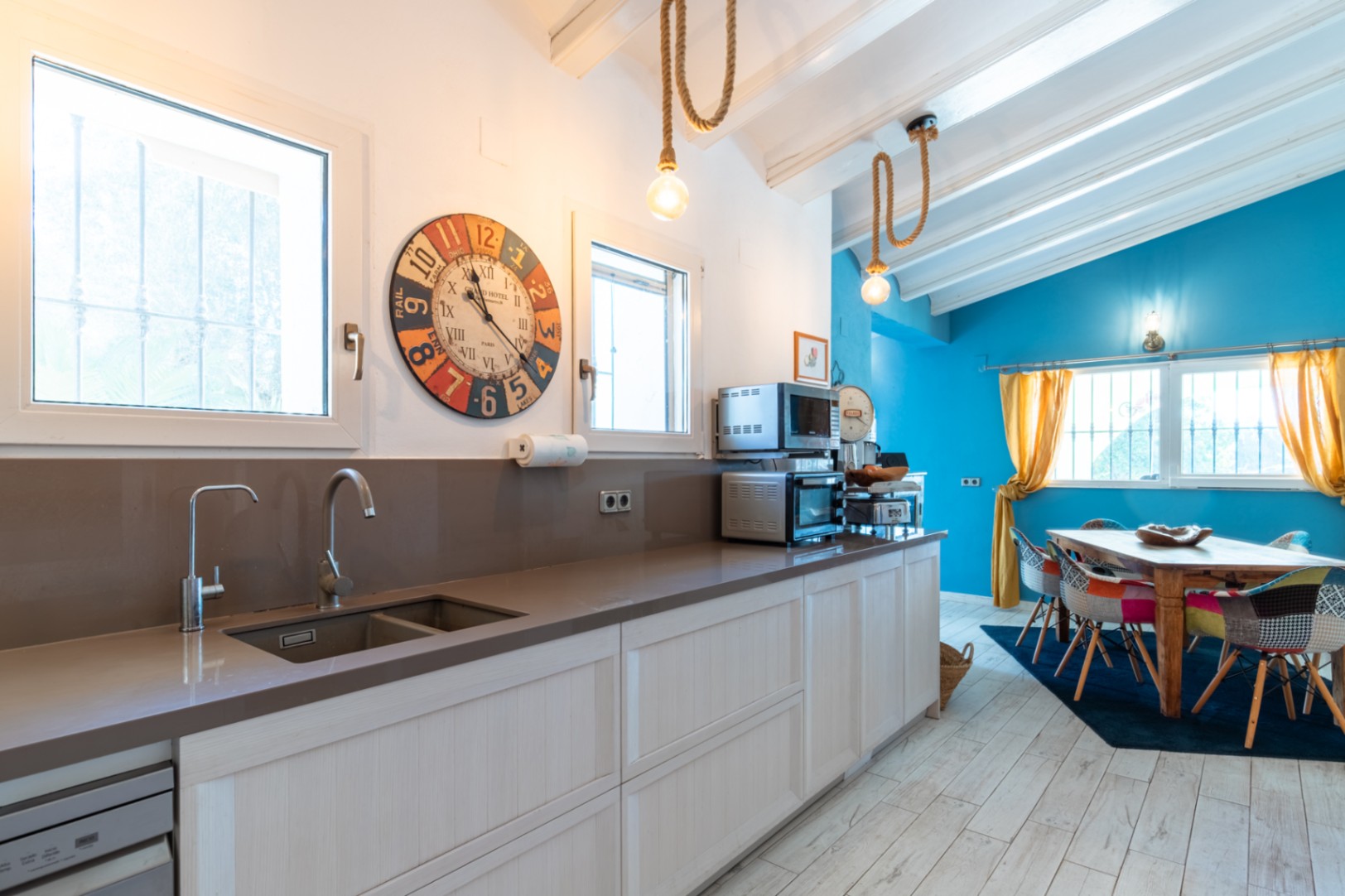 Moderne Küche mit Quarz-Arbeitsplatte, offen zu einem hellen Essbereich mit blauer Akzentwand, weißen Deckenbalken und rustikalen Seil-Pendelleuchten.