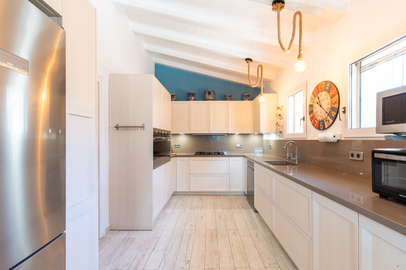 Moderne Küche mit Steinarbeitsplatten, Edelstahlelementen und weißer Balkendecke. Dekorative Seilleuchten und ein blauer Farbakzent prägen das helle Küchendesign.