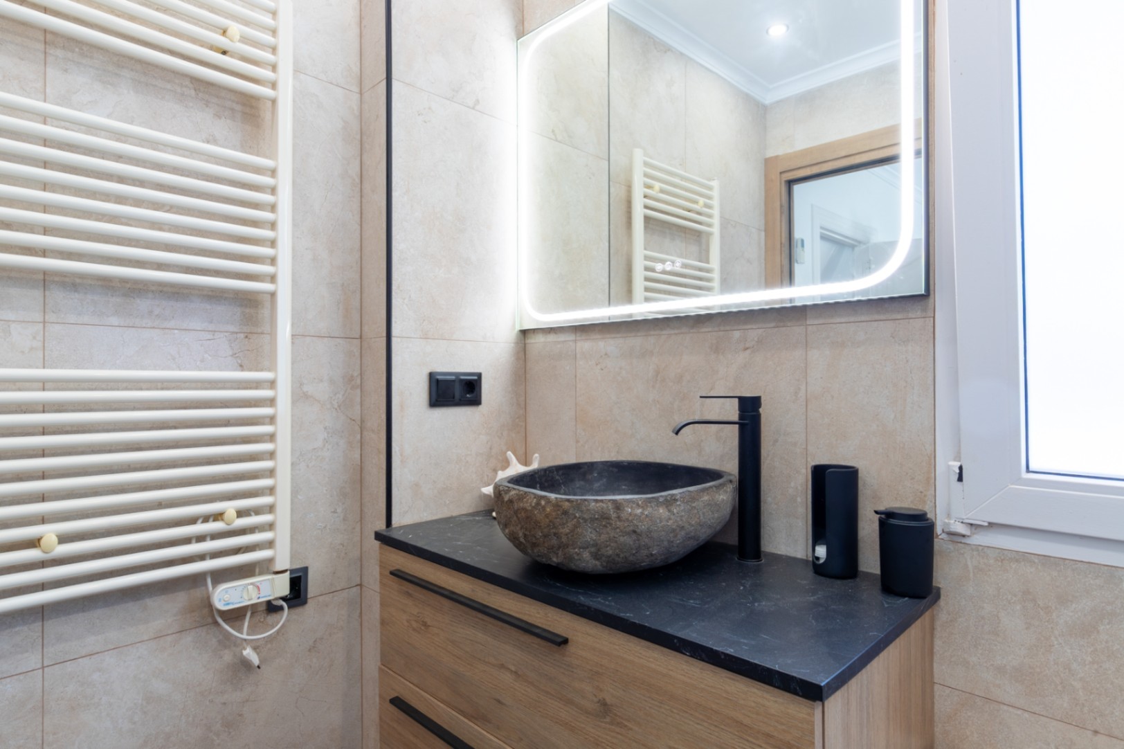 Modernes Badezimmer mit Naturstein-Aufsatzwaschbecken auf schwarzer Arbeitsplatte und Holzunterschrank. Mit LED-hinterleuchtetem Spiegel und weißem Handtuchheizkörper.