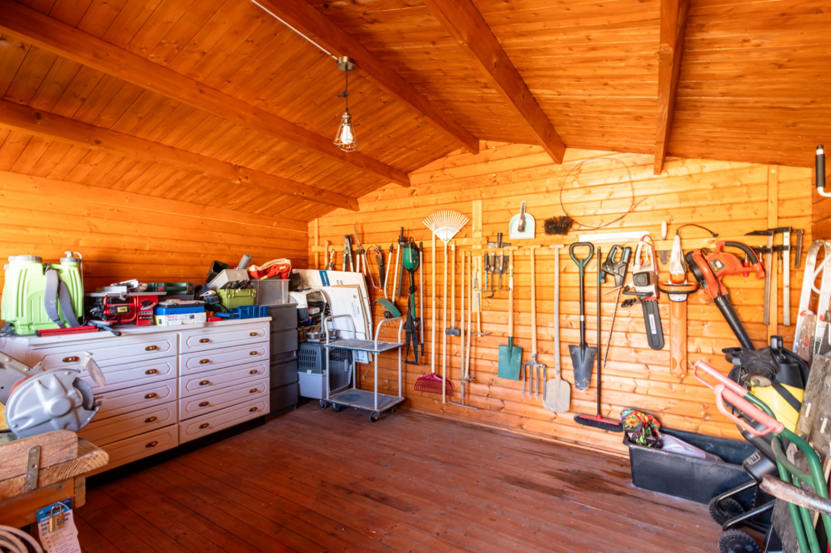 Organisiertes Gartenhaus aus Holz mit Wandhalterungen für Werkzeuge, einer weißen Kommode und sichtbarer Balkenkonstruktion.
