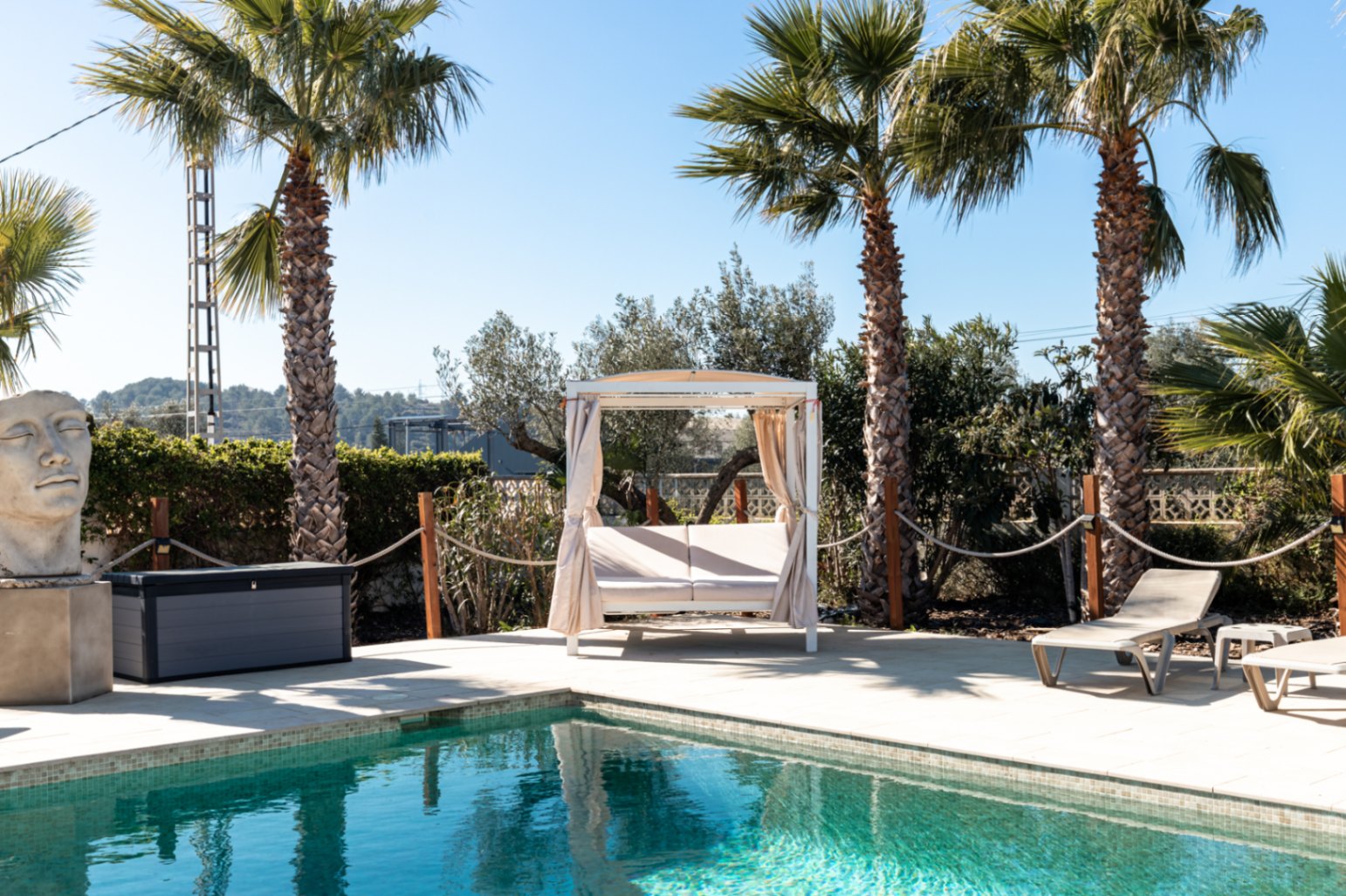 Poolbereich mit Natursteinterrasse, balinesischem Tagesbett mit Vorhängen, Sonnenliegen und mediterraner Bepflanzung mit hohen Palmen.