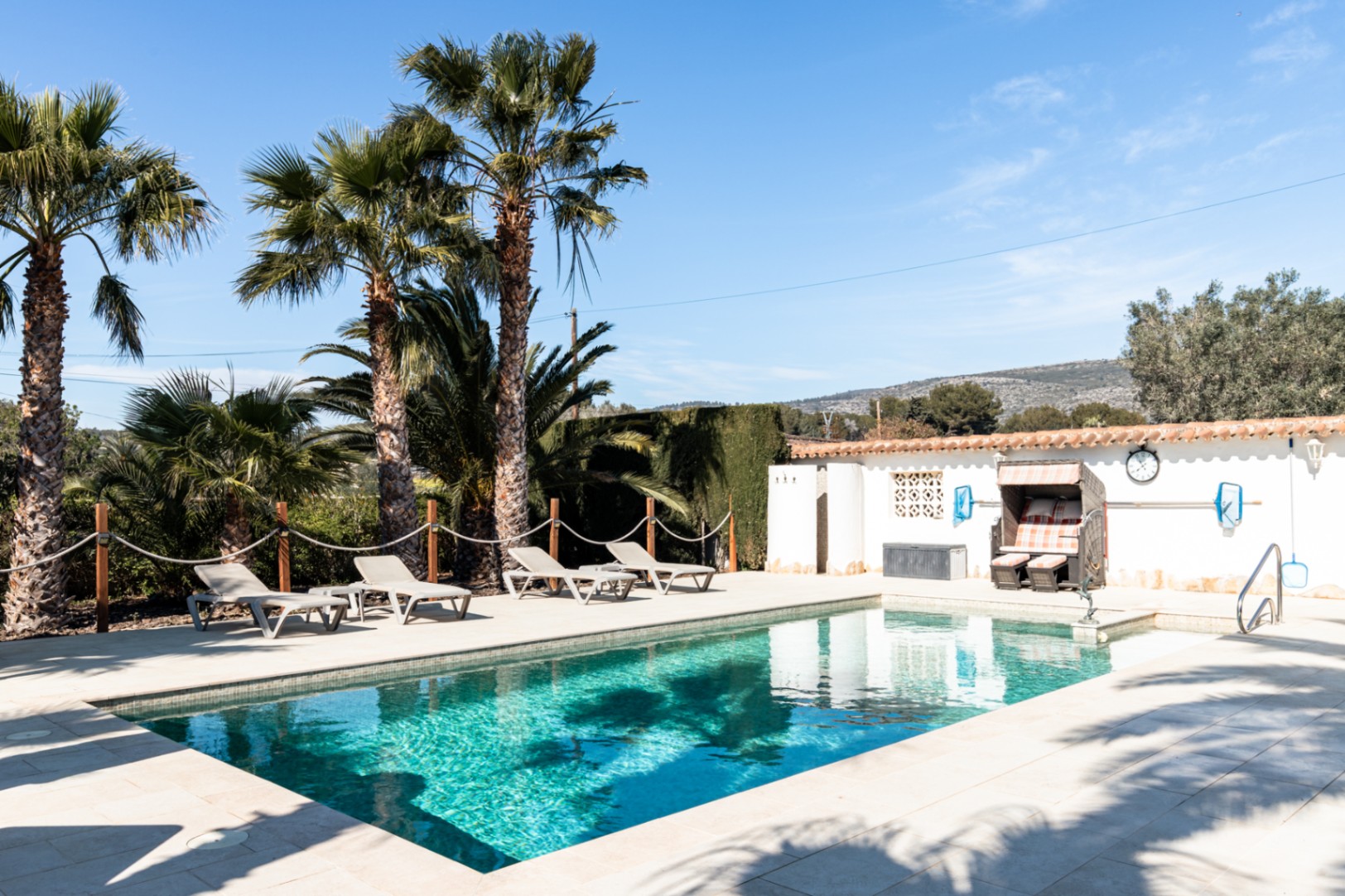 Privater 9,5m x 5m Pool mit heller Steinterrasse, Sonnenliegen und Palmen, ergänzt durch einen Strandkorb und Blick auf die umliegende Hügellandschaft.
