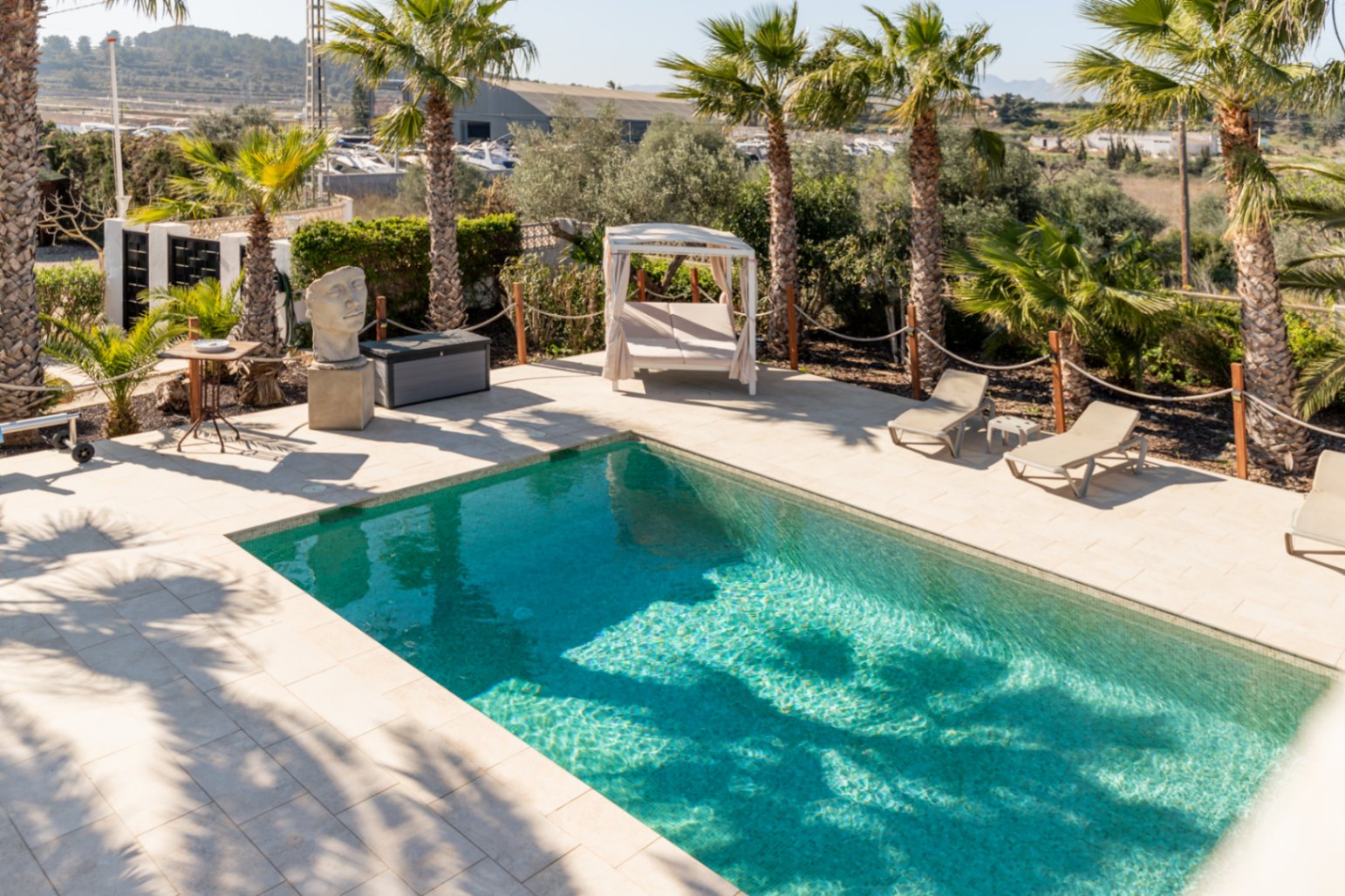Rechteckiger Swimmingpool mit heller Steinterrasse, Sonnenliegen und einem balinesischen Daybed, umgeben von Palmen und dekorativen Gartenelementen.