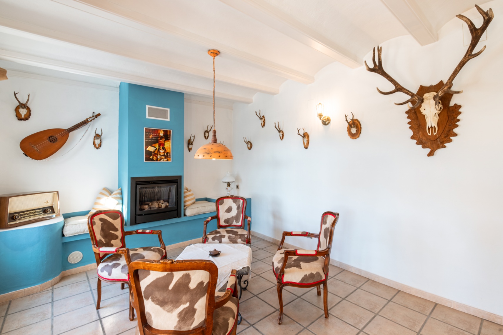 Rustikales Wohnzimmer mit Gaskamin in blauer Akzentwand, Sesseln mit Kuhfell-Polsterung und weißen Balkendecken mit jagdlicher Dekoration.