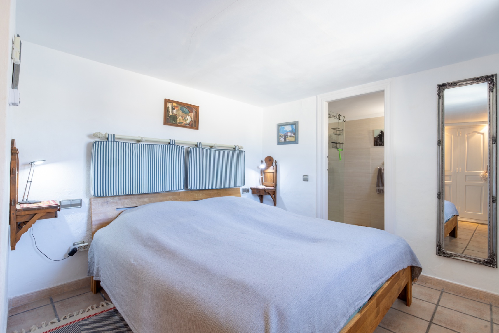 Schlafzimmer mit Holzbettrahmen, gestreiften Kopfteilkissen und wandmontierten Nachttischen. Mit direktem Zugang zum En-Suite-Badezimmer und einem großen verzierten Wandspiegel.