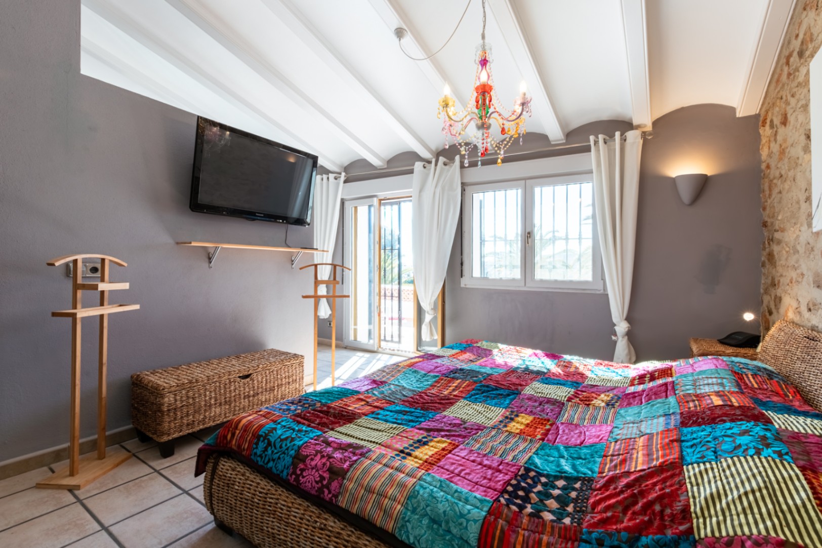 Schlafzimmer mit Natursteinwand, weißer Balkendecke und Korbbett. Ausgestattet mit einem bunten Kronleuchter und direktem Zugang zum Außenbereich.