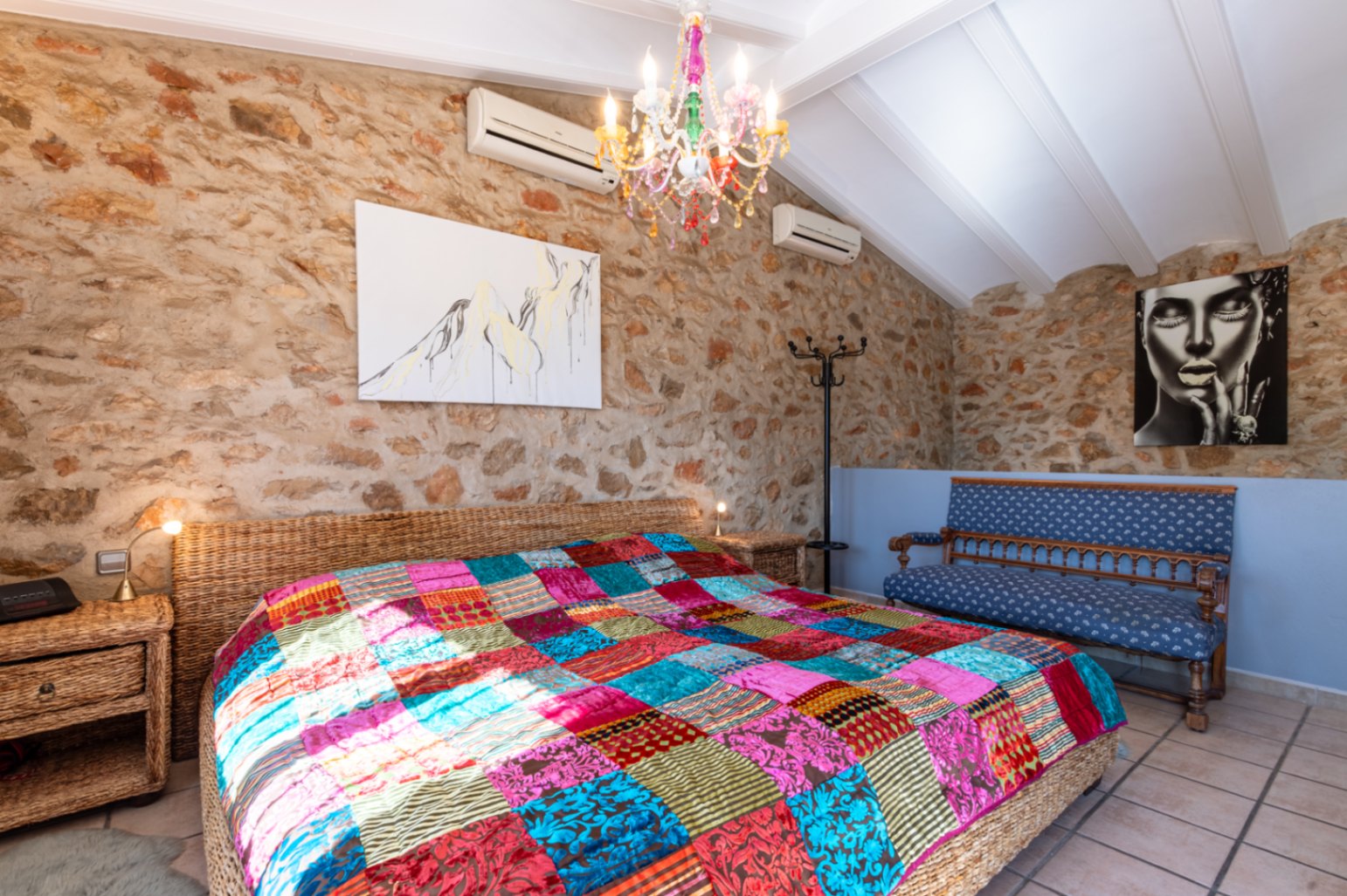 Schlafzimmer mit Natursteinwänden, weißer Balkendecke und einem Korbbett mit bunter Patchwork-Decke unter einem farbenfrohen Kristallleuchter.