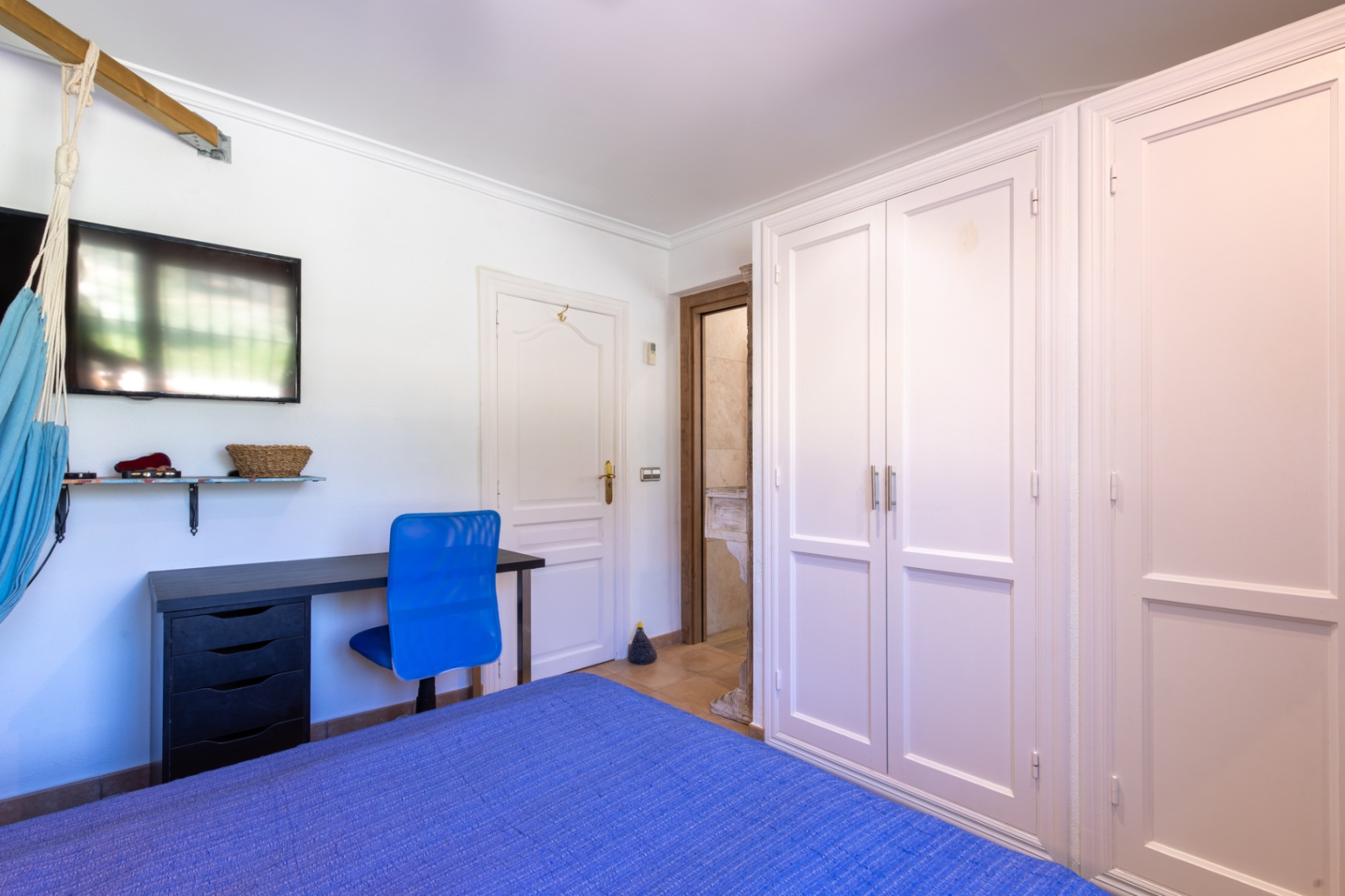 Schlafzimmer mit weißen Einbauschränken, Arbeitsbereich mit blauem Stuhl und Zugang zu einem En-Suite-Badezimmer mit Natursteinelementen.