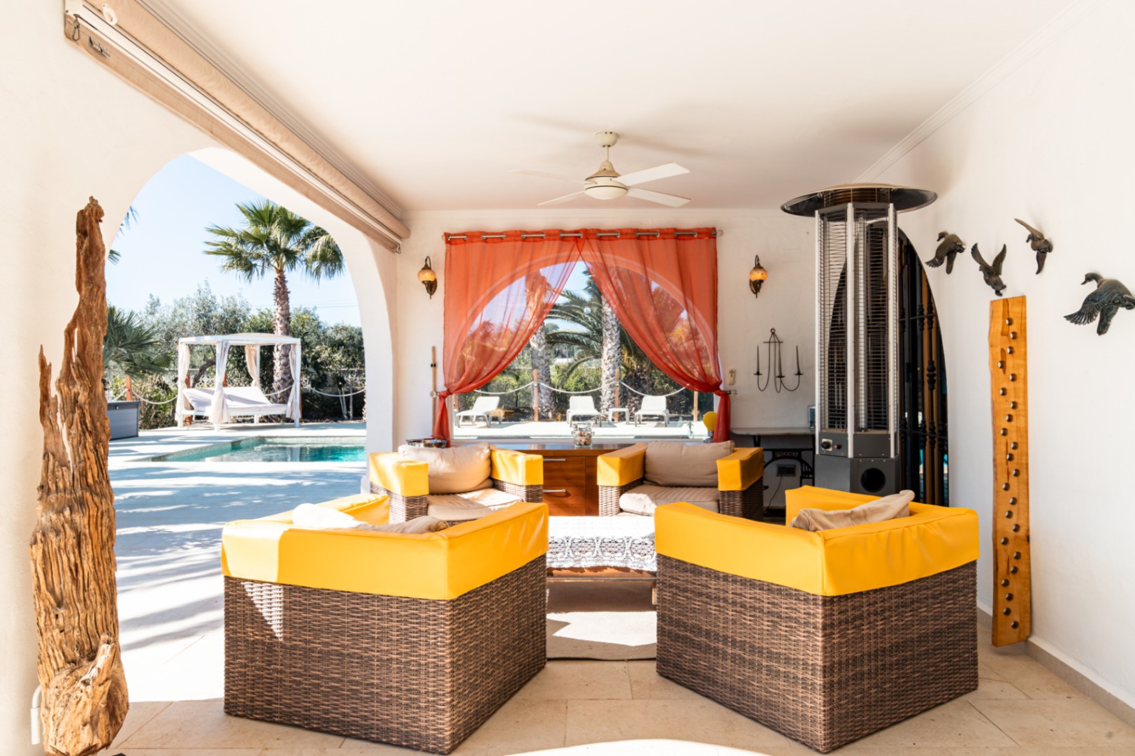 Überdachte Terrasse mit Lounge-Sesseln aus Korbgeflecht, Deckenventilator und Heizstrahler, mit Blick auf den Poolbereich und den mediterranen Garten.