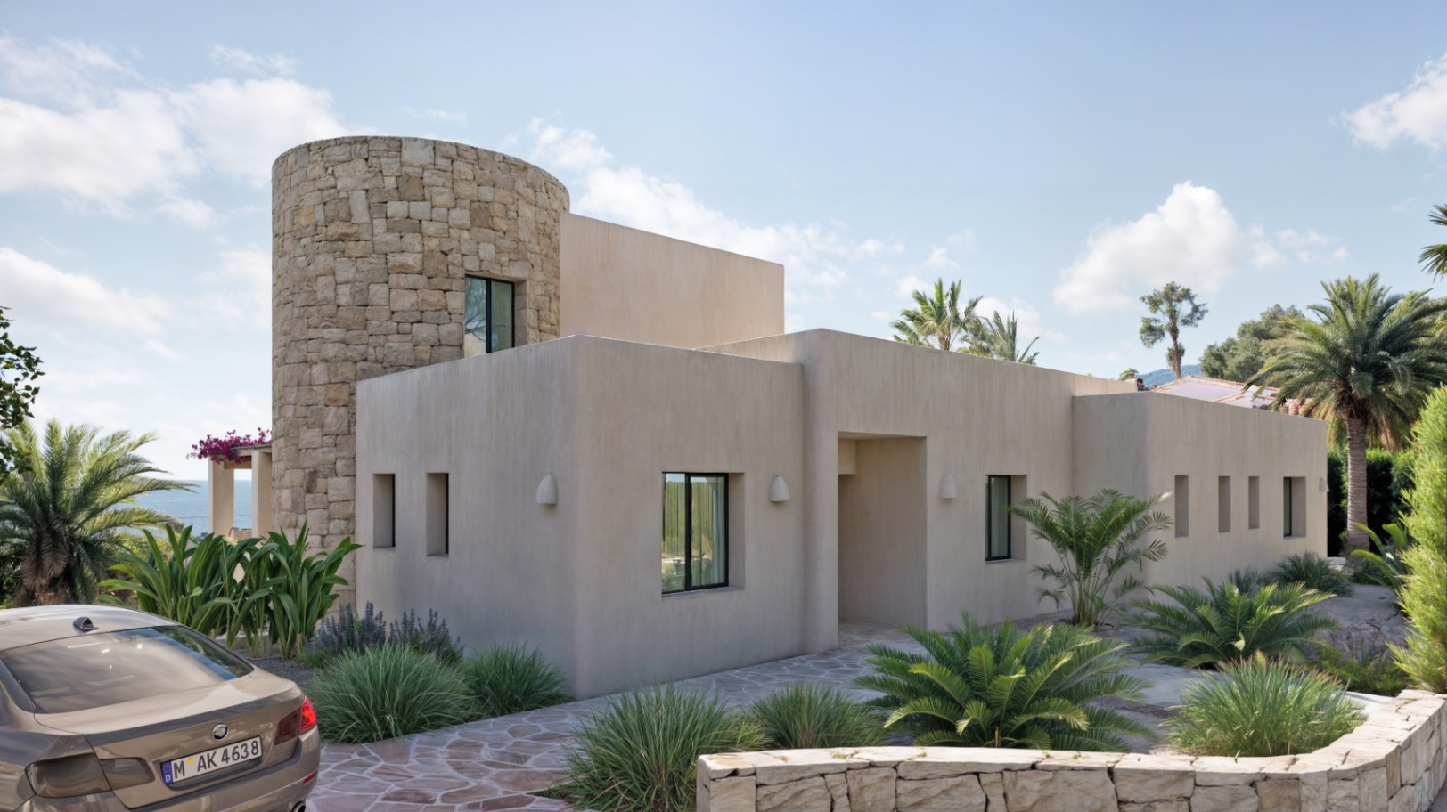 Außenansicht im Ibiza-Stil mit einem Rundturm aus Naturstein, schwarzen Aluminiumfenstern und mediterraner Bepflanzung auf einem gepflasterten Grundstück.