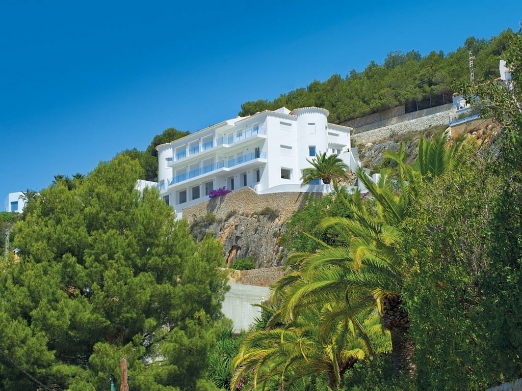 Außenansicht der weißen, in den Fels gebauten Villa mit mehreren Terrassenebenen, Glasgeländern und üppiger mediterraner Bepflanzung aus Pinien und Palmen.