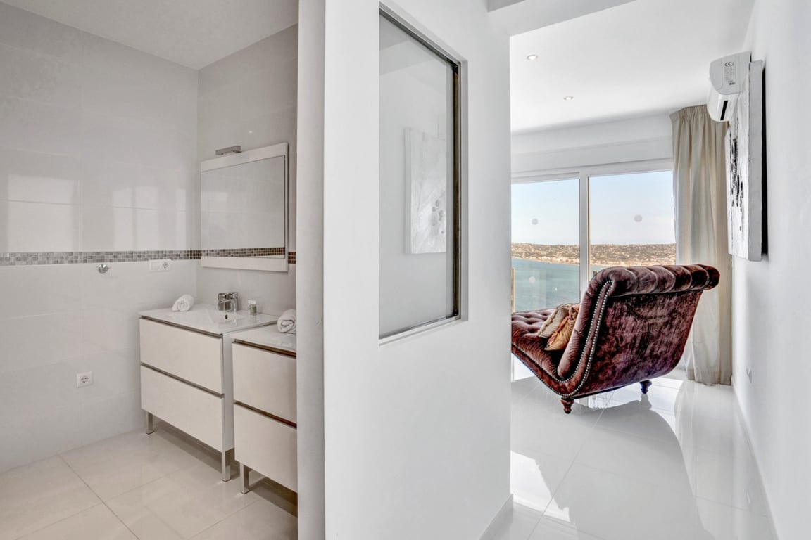 En-Suite-Badezimmer mit Doppelwaschtisch und Mosaikdetails, das an einen hellen Wohnbereich mit Samt-Chaiselongue und Meerblick grenzt.