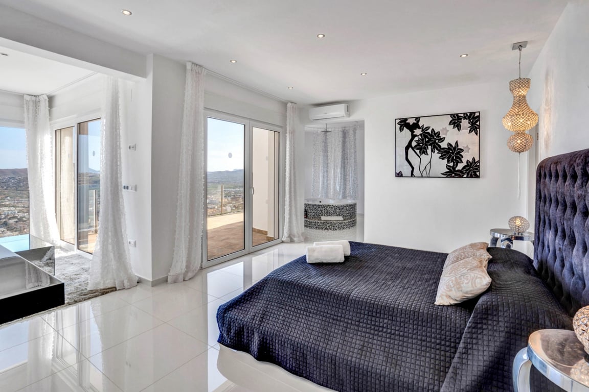 Hauptschlafzimmer mit gepolstertem Kopfteil, glänzenden Fliesen und direktem Terrassenzugang. Ein integrierter Mosaik-Whirlpool bietet Ausblick auf die Bucht.