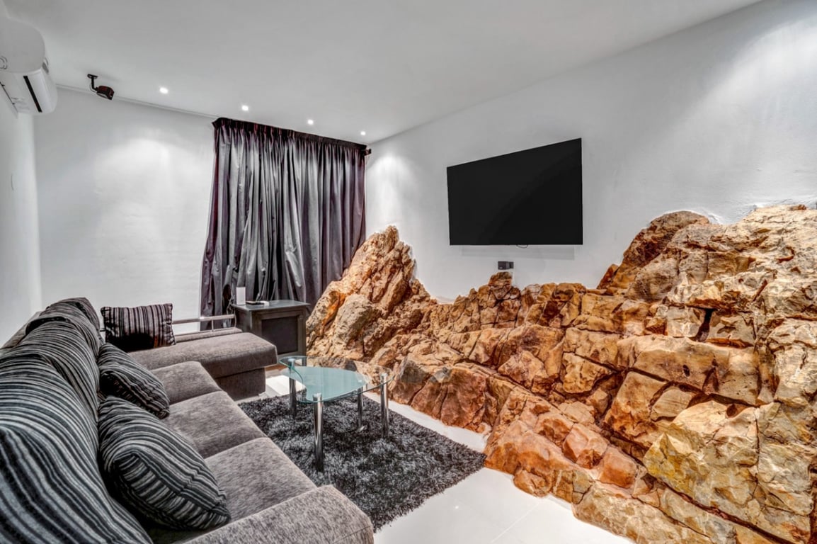 Heimkino-Raum mit in die Wand integriertem Naturfels, grauem Sofa, Glastisch und wandmontiertem Fernseher mit Einbaustrahlern.