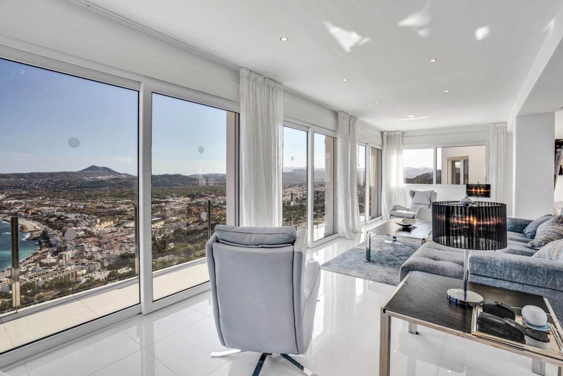 Heller Wohnbereich mit bodentiefen Fenstern, Panoramablick auf die Bucht von Jávea und die Berge, ausgestattet mit weißen Glanzböden und moderner Beleuchtung.