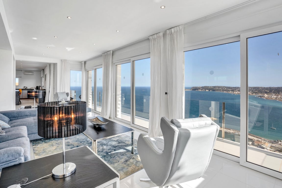 Heller Wohnbereich mit bodentiefen Glasschiebetüren und Panoramablick auf das Mittelmeer. Die Ausstattung umfasst moderne Sitzmöbel, Einbaustrahler und helle Bodenfliesen.