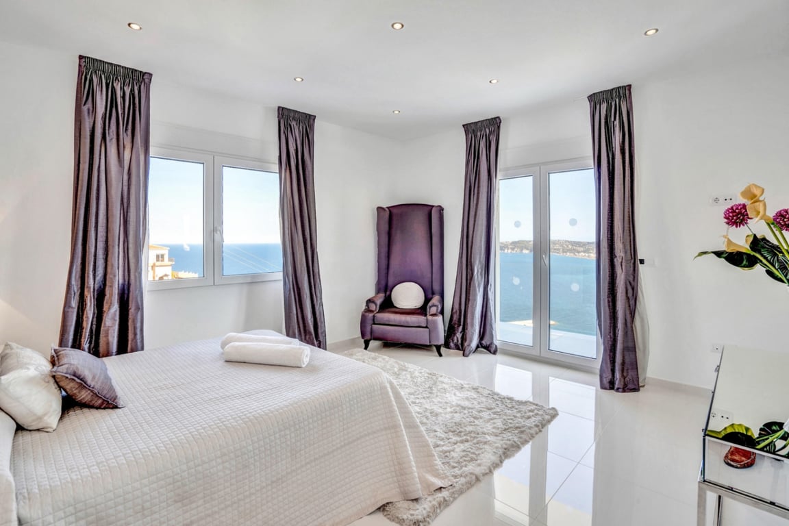Helles Schlafzimmer mit violettem Samtsessel, weißen Fliesenböden und großen Fenstern mit Panoramablick auf das Mittelmeer in Jávea.