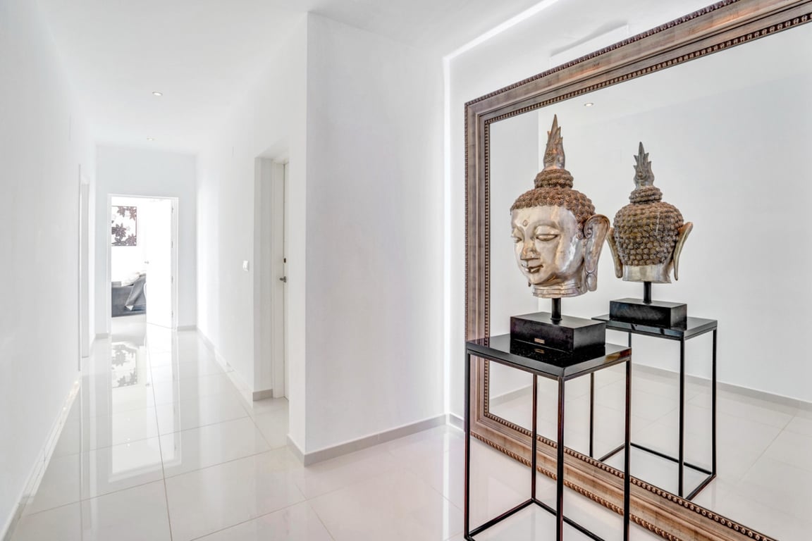 Minimalistischer Flur mit poliertem Fliesenboden und Einbaustrahlern. Ein großer gerahmter Spiegel reflektiert eine silberne Buddha-Skulptur auf einem schwarzen Sockel.